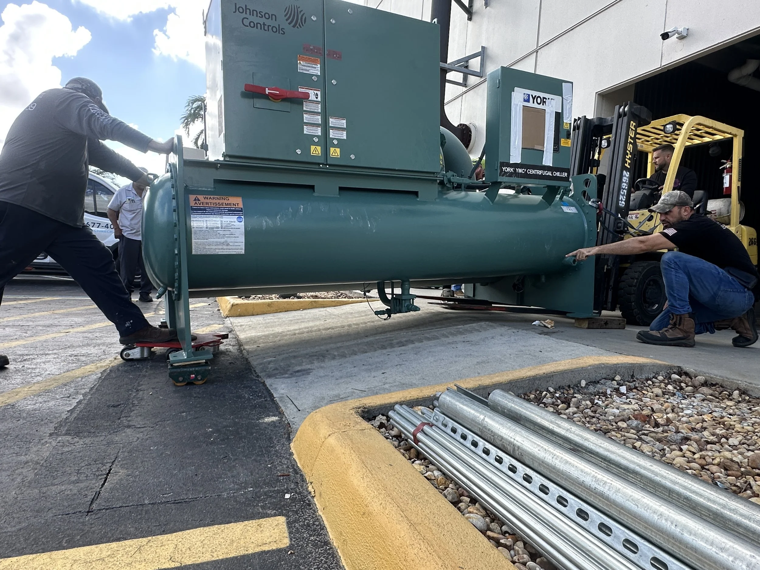 Workers installing or repairing a large industrial cooling unit outside a building, with a forklift helping position the equipment and pipes laid on the ground nearby.