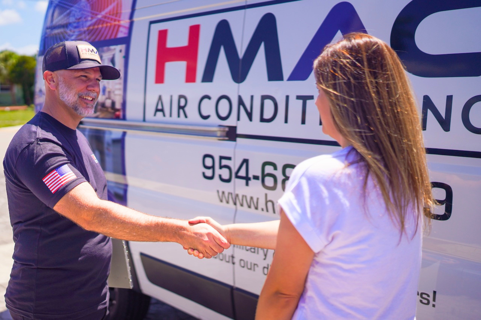 A man with a beard wearing a cap and a navy T-shirt with an American flag patch on the sleeve shakes hands with a woman with long, light brown hair, in front of a moving van with the company name "HMA Air Conditioning" visible on it.