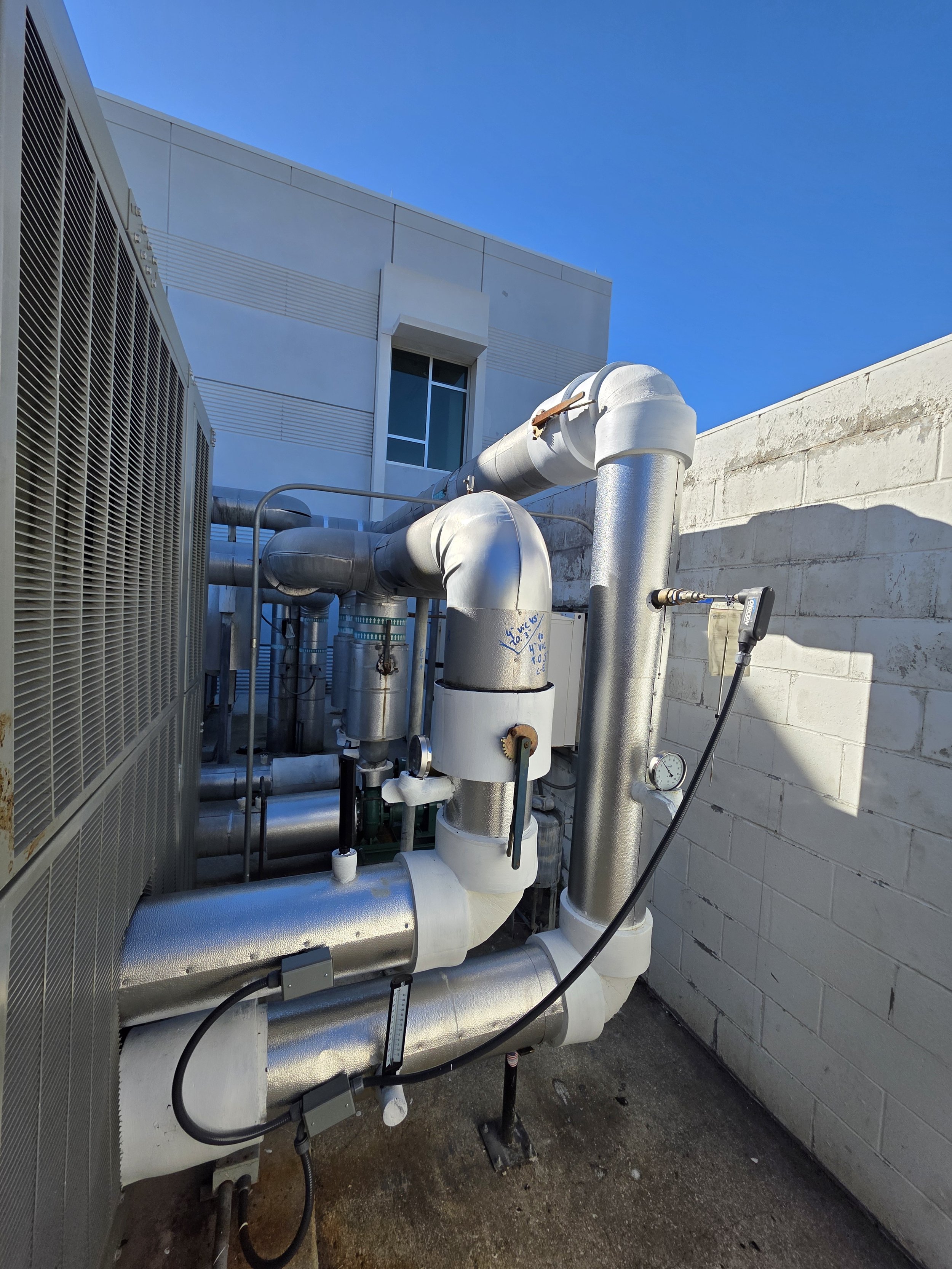 On a rooftop, there are large white HVAC pipes, a gray metal unit, and a white cinder block wall under a clear blue sky.