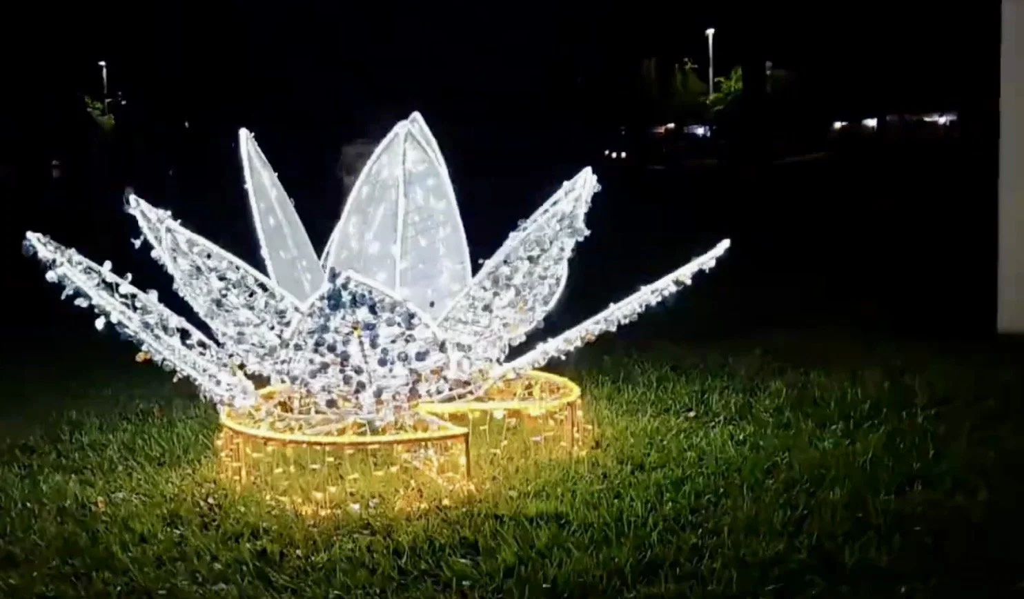 Illuminated large decorative lotus flower sculpture with white lights, placed on grass at night.