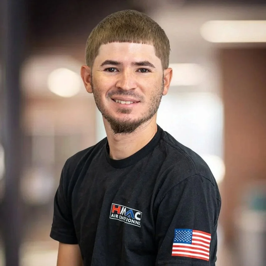 Young man with brown hair and a beard smiling, wearing a black t-shirt with HMC Air Conditioning logo and American flag patches on the sleeves, standing against a plain white background.