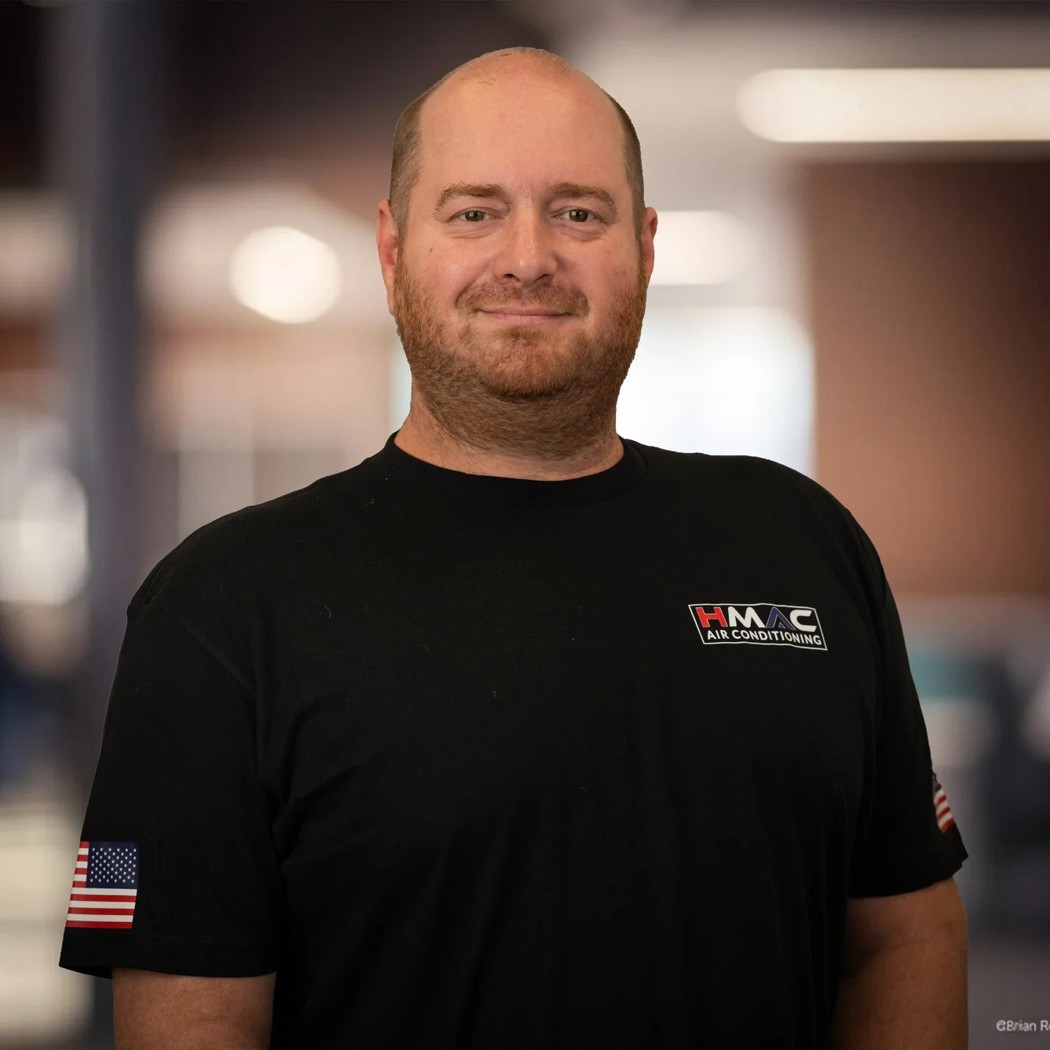 A man with a beard and short hair, wearing a black t-shirt with American flag patches on the sleeves and a logo that says "HMAC Air Conditioning," standing in an indoor setting with blurred background lighting.