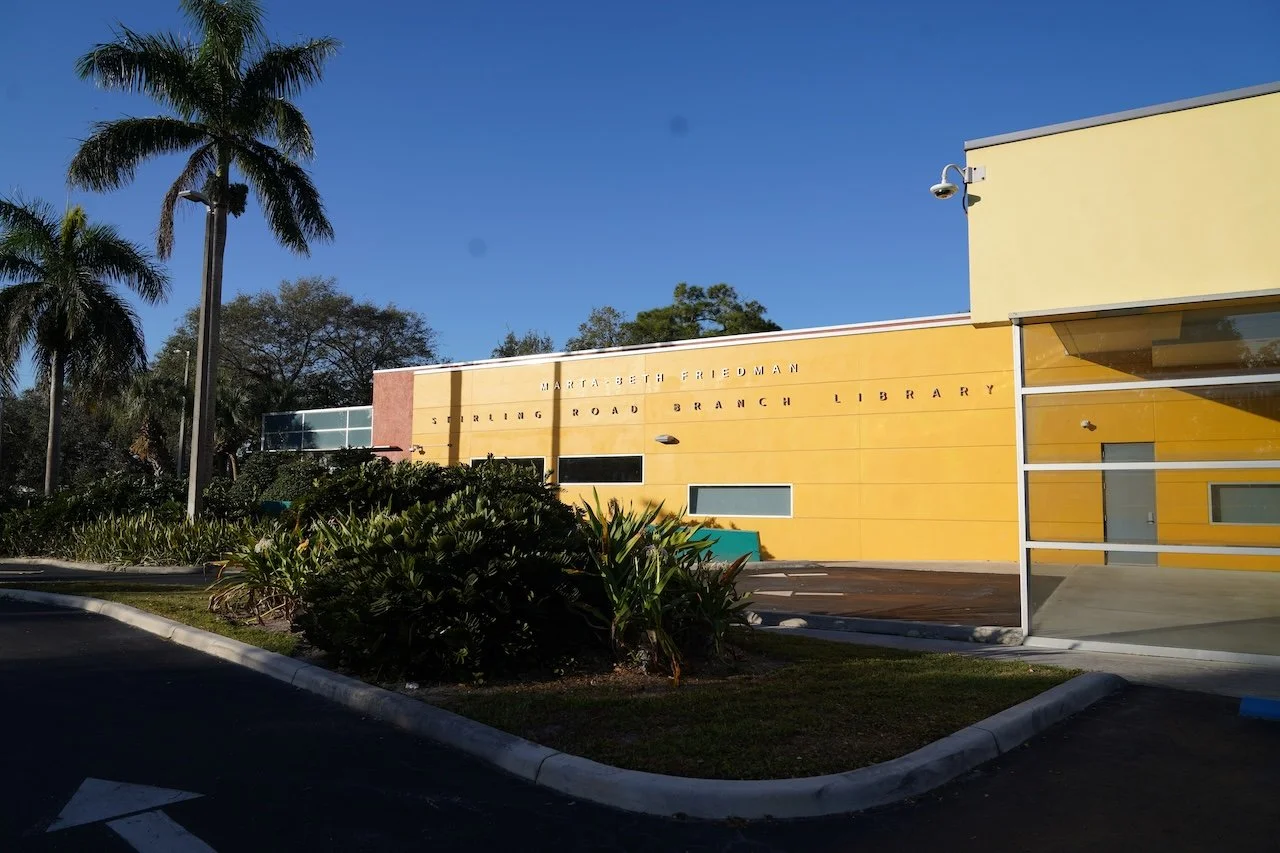 Exterior of a yellow building labeled 'Marta Beth Friedman Shirley Road Branch Library' with tropical trees and plants, parking lot, and a clear blue sky.