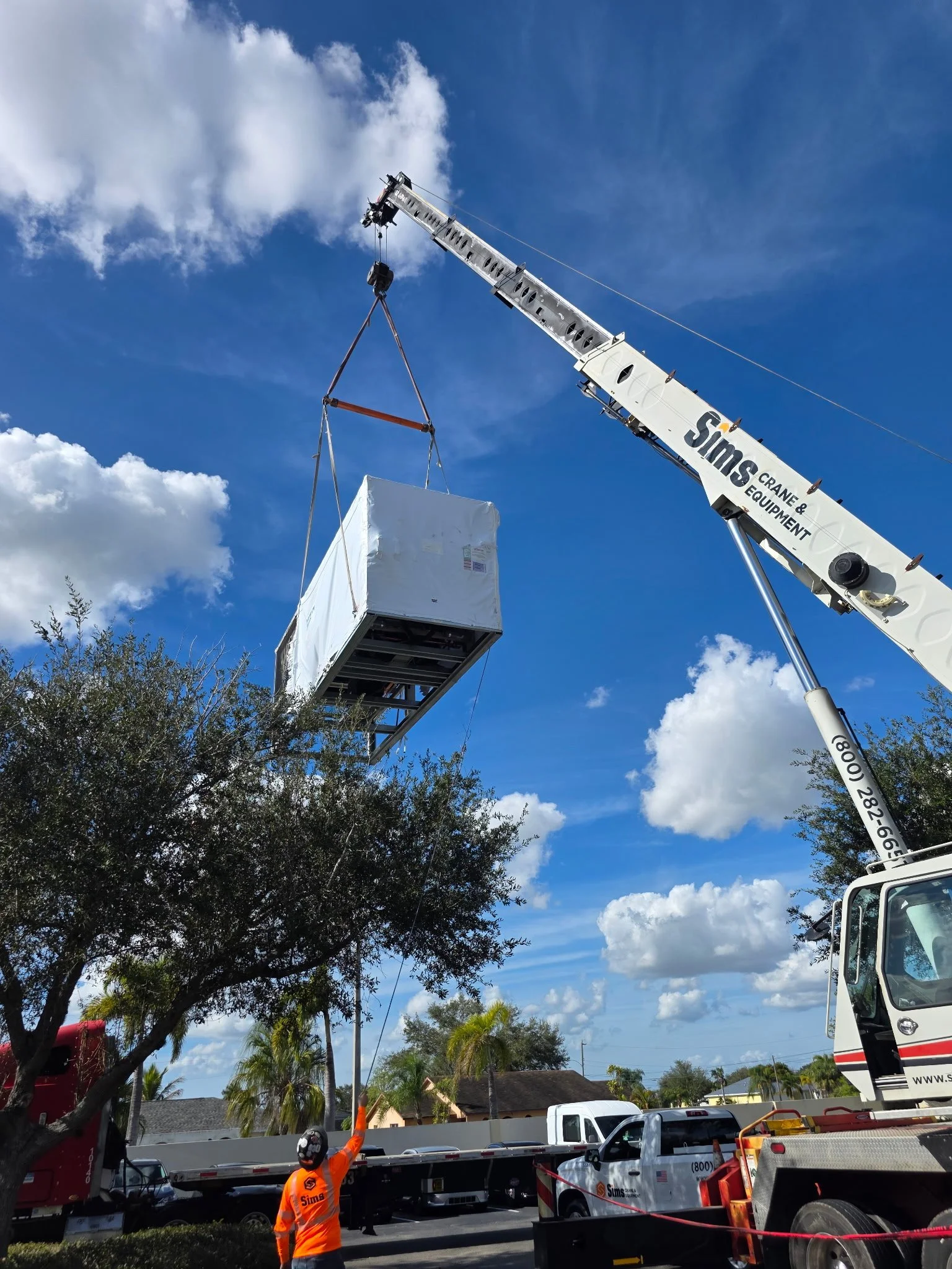 A crane lifting a large white block in a residential area with palm trees, blue sky, and white clouds in the background.