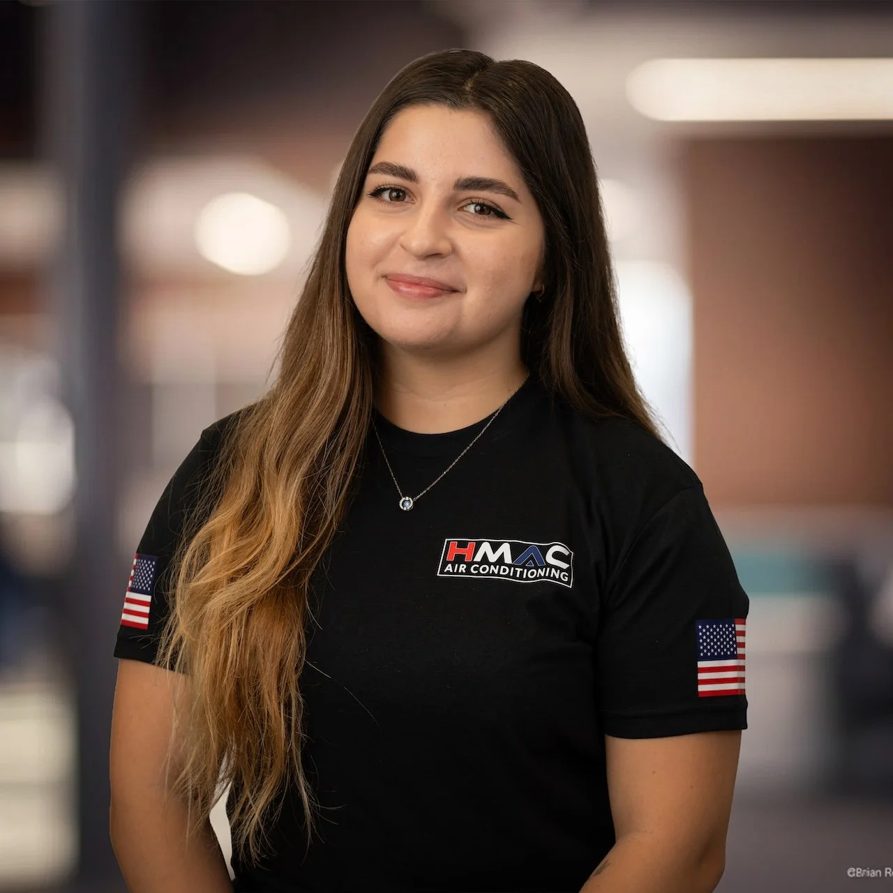 A young woman with long brown hair wearing a black HVAC company shirt with American flags on the sleeves, smiling at the camera.