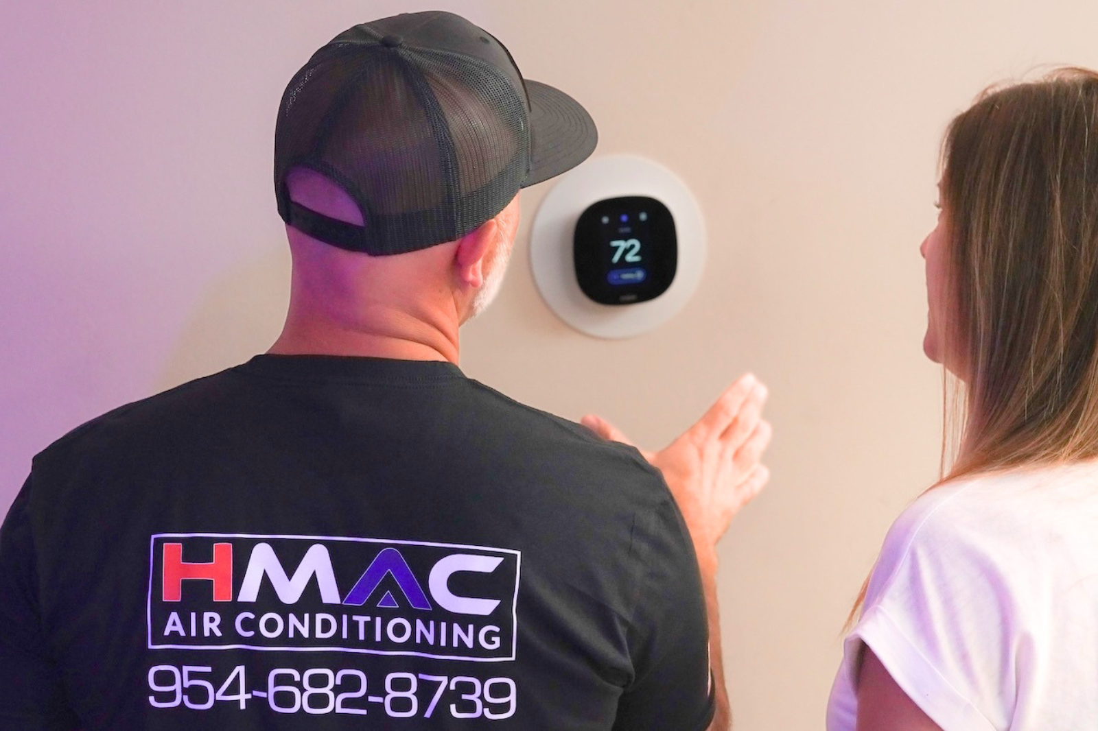 A man wearing a black cap and a black t-shirt with the logo 'HMAC Air Conditioning' and phone number on the back, showing a woman with long hair in a light-colored shirt, as he discusses the thermostat displaying a temperature of 72 degrees Fahrenheit on the wall.
