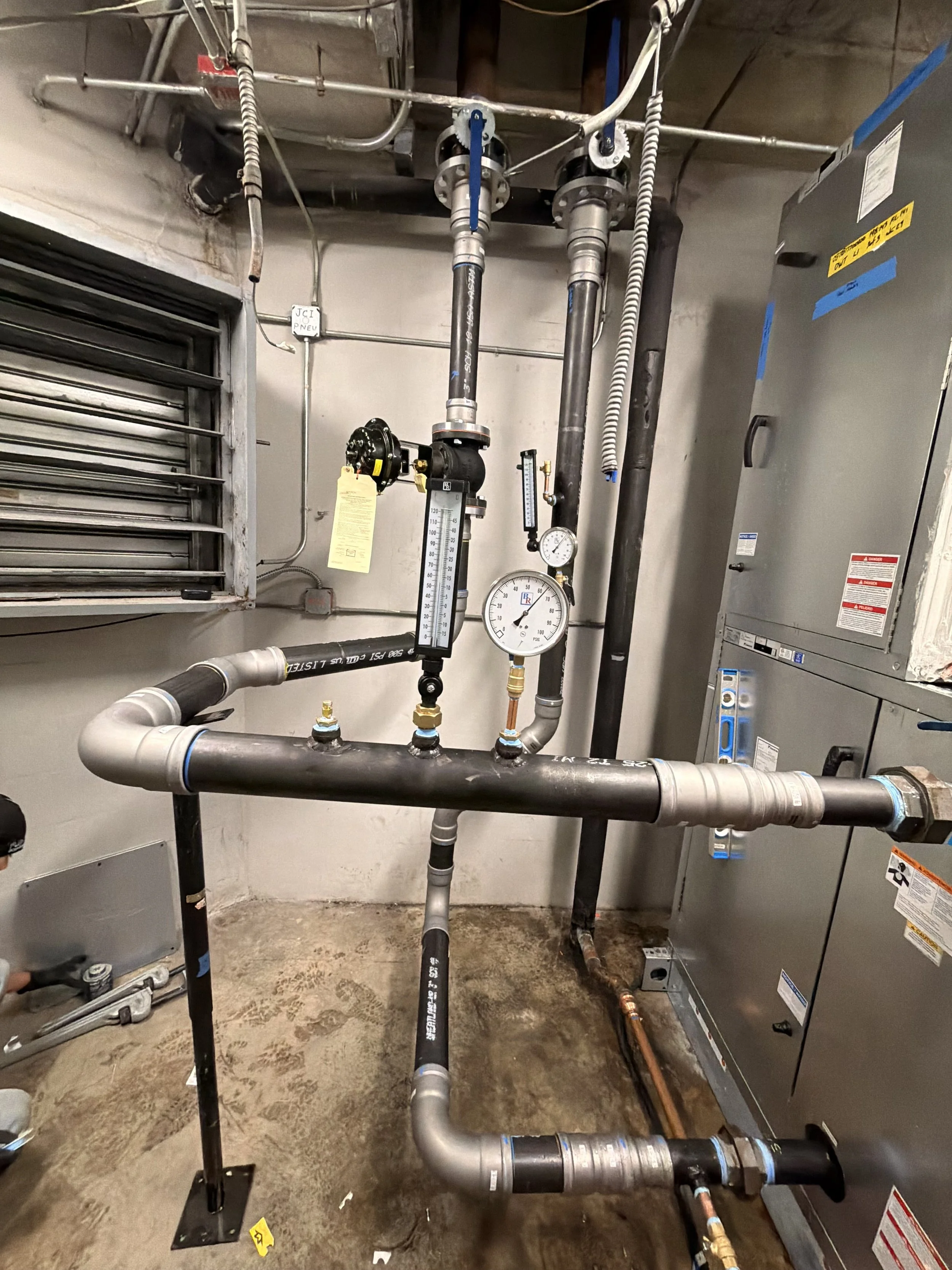 The image shows a utility room with a complex system of pipes, gauges, and equipment. There are black and gray pipes, a pressure gauge, and a thermostat. A large gray electrical panel is on the right side, and a small window with metal louvers is on the left side. The concrete floor is visible, and some tools are on the ground.
