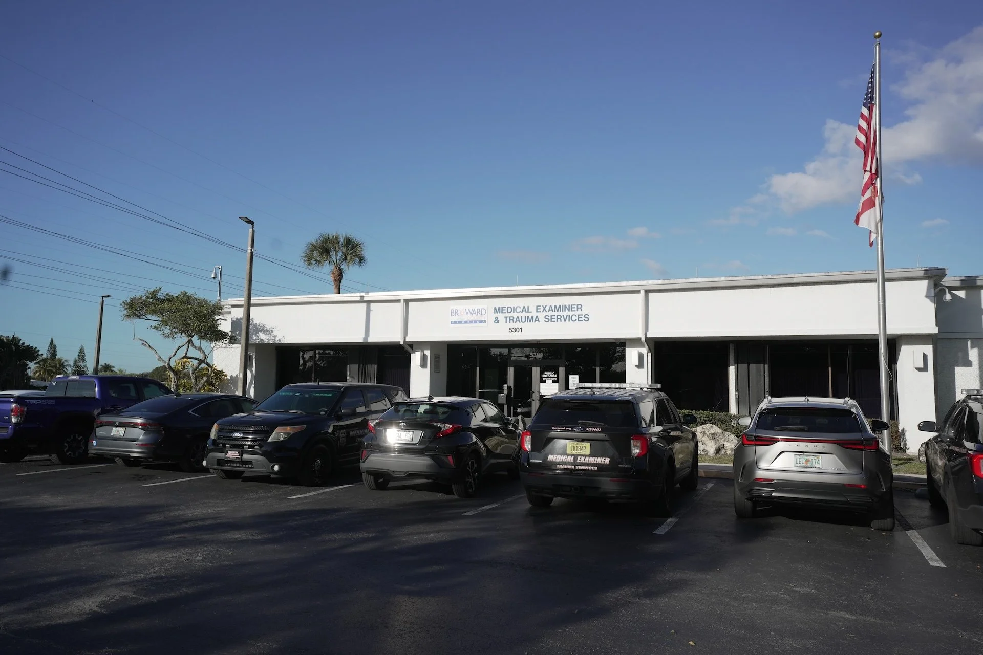 Parking lot in front of a white building with a sign that reads 'BR WARD Medical Examiner & Trauma Services'. Several cars are parked, and there are American flags, palm trees, and utility poles visible in the background under a partly cloudy sky.