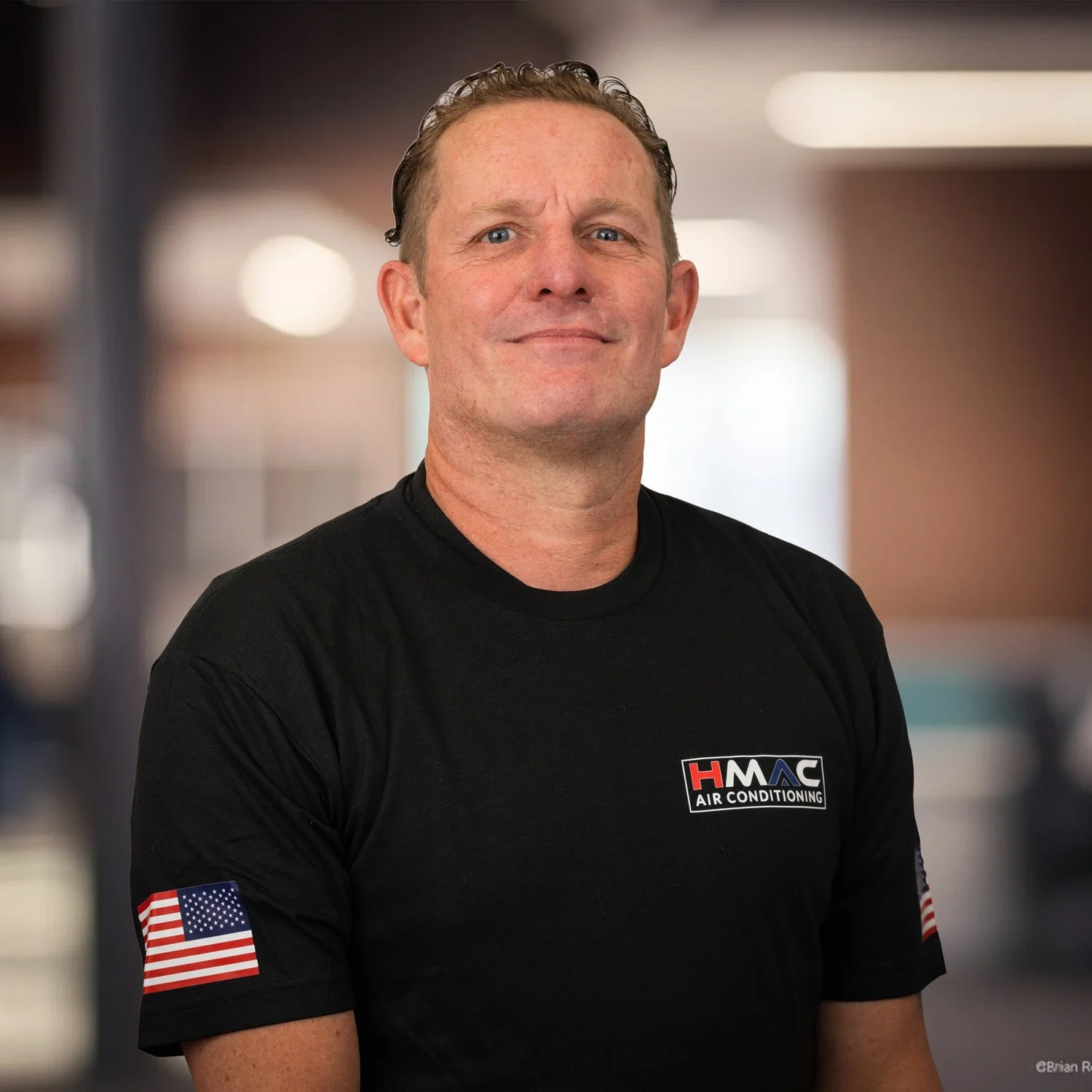 A man in a black shirt with an American flag patch on his sleeve and a logo for HVAC air conditioning on his chest, standing indoors with a blurred background.