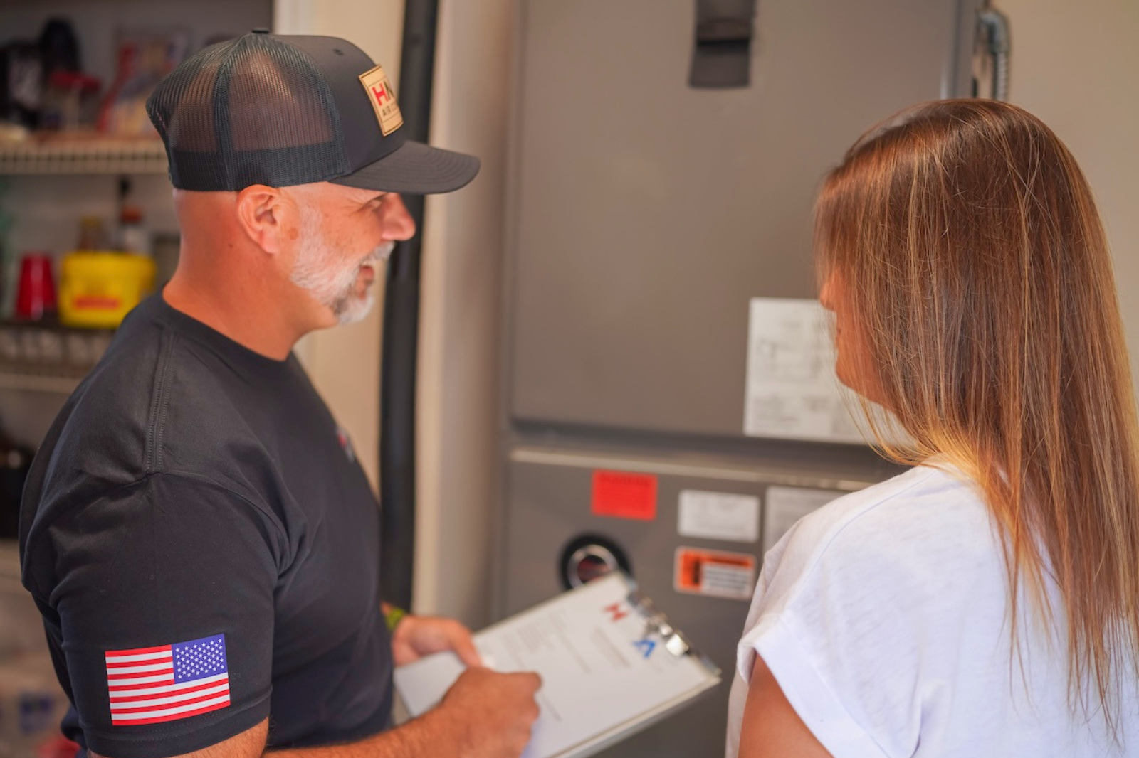 A man with gray hair and a beard, wearing a black cap and black shirt with American flag patches, is holding a clipboard and talking to a woman with long brown hair, in front of a gray appliance in a kitchen or utility room.