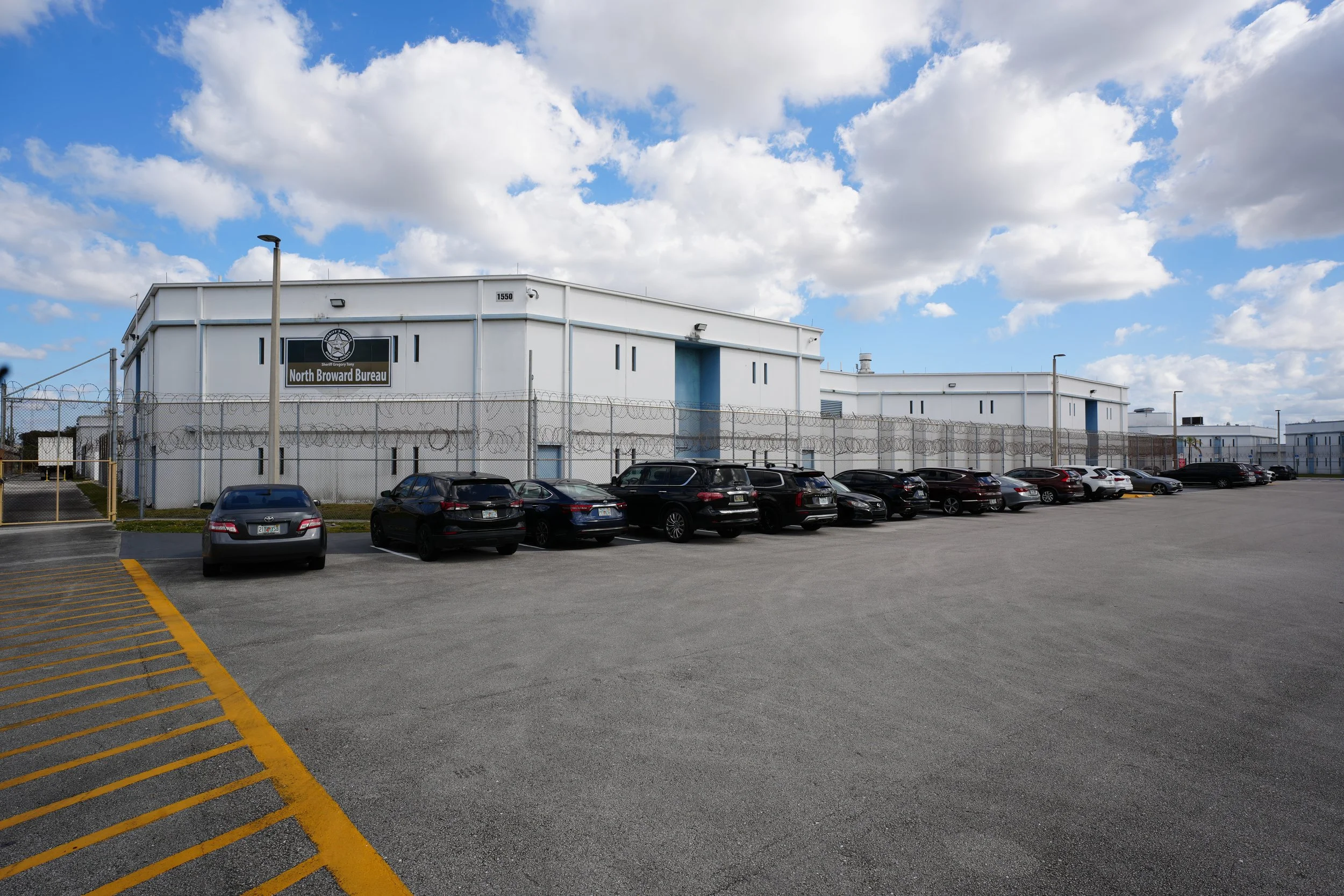 A parking lot in front of a white building labeled 'North Broward Bureau' with a fence and multiple parked cars under a partly cloudy sky.