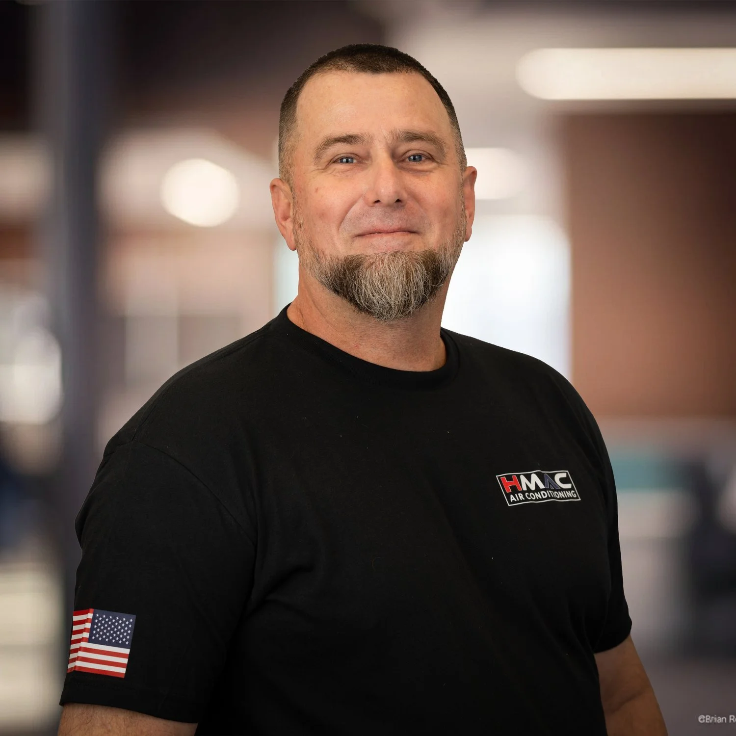 A man with short hair, a beard, and blue eyes, wearing a black t-shirt with a logo that says "HMC Air Conditioning" on the chest, standing in a well-lit indoor setting with a blurred background.