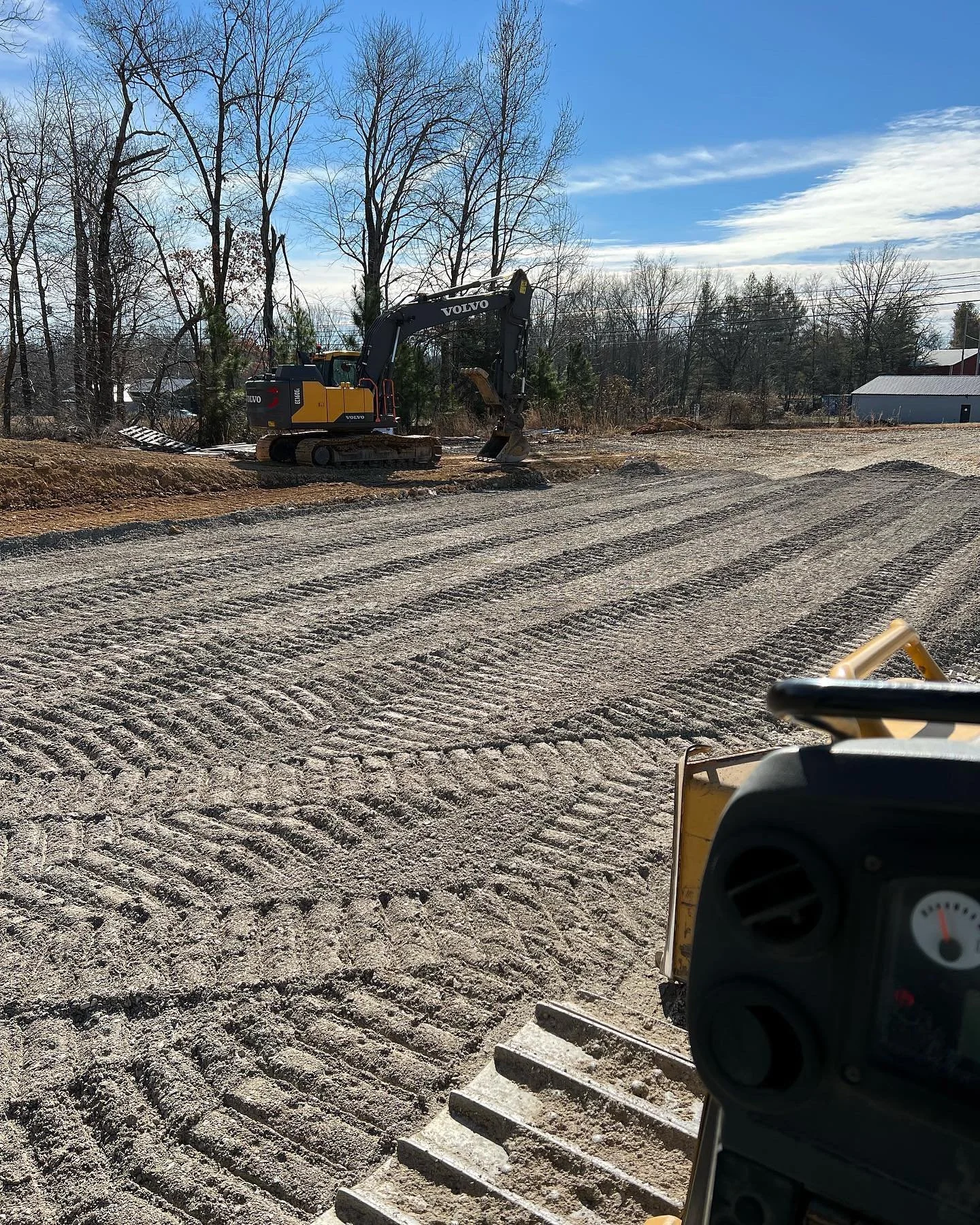 Construction site with a Volvo excavator on a dirt ground, some geometrics patterns in the earth, and trees with bare branches in the background under a partly cloudy sky.