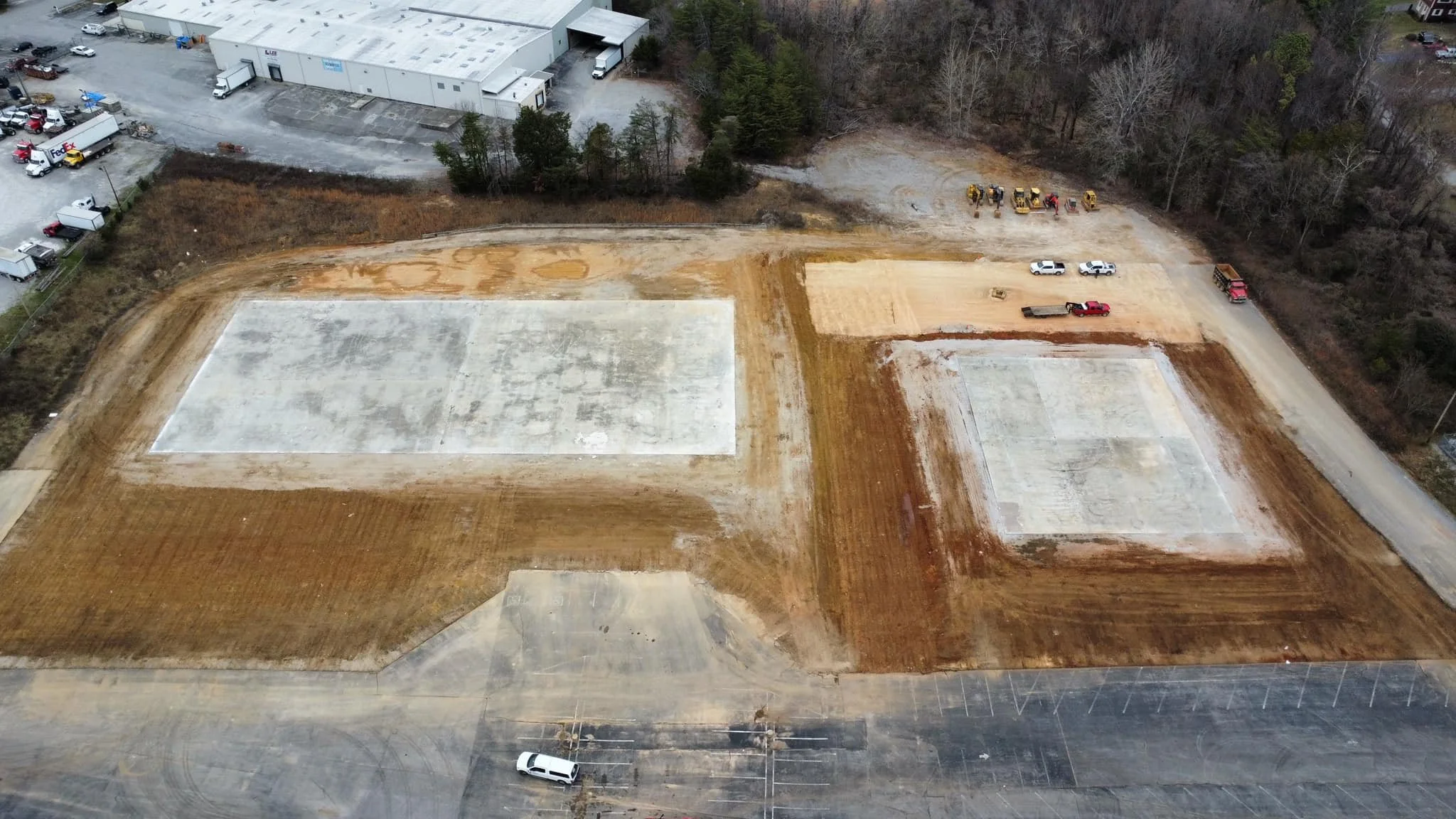 Aerial view of a construction site with cleared and leveled ground for future buildings, parked vehicles, and construction equipment, surrounded by trees and adjacent to industrial buildings.