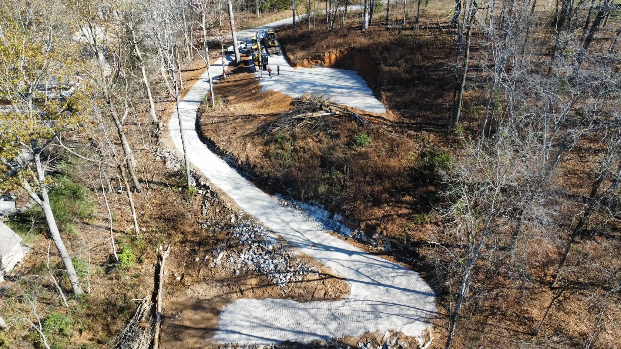 An aerial view of a construction site on a hillside with trees, where a section of road is being replaced with concrete. Construction equipment and workers are present, and some trees have been cut down.