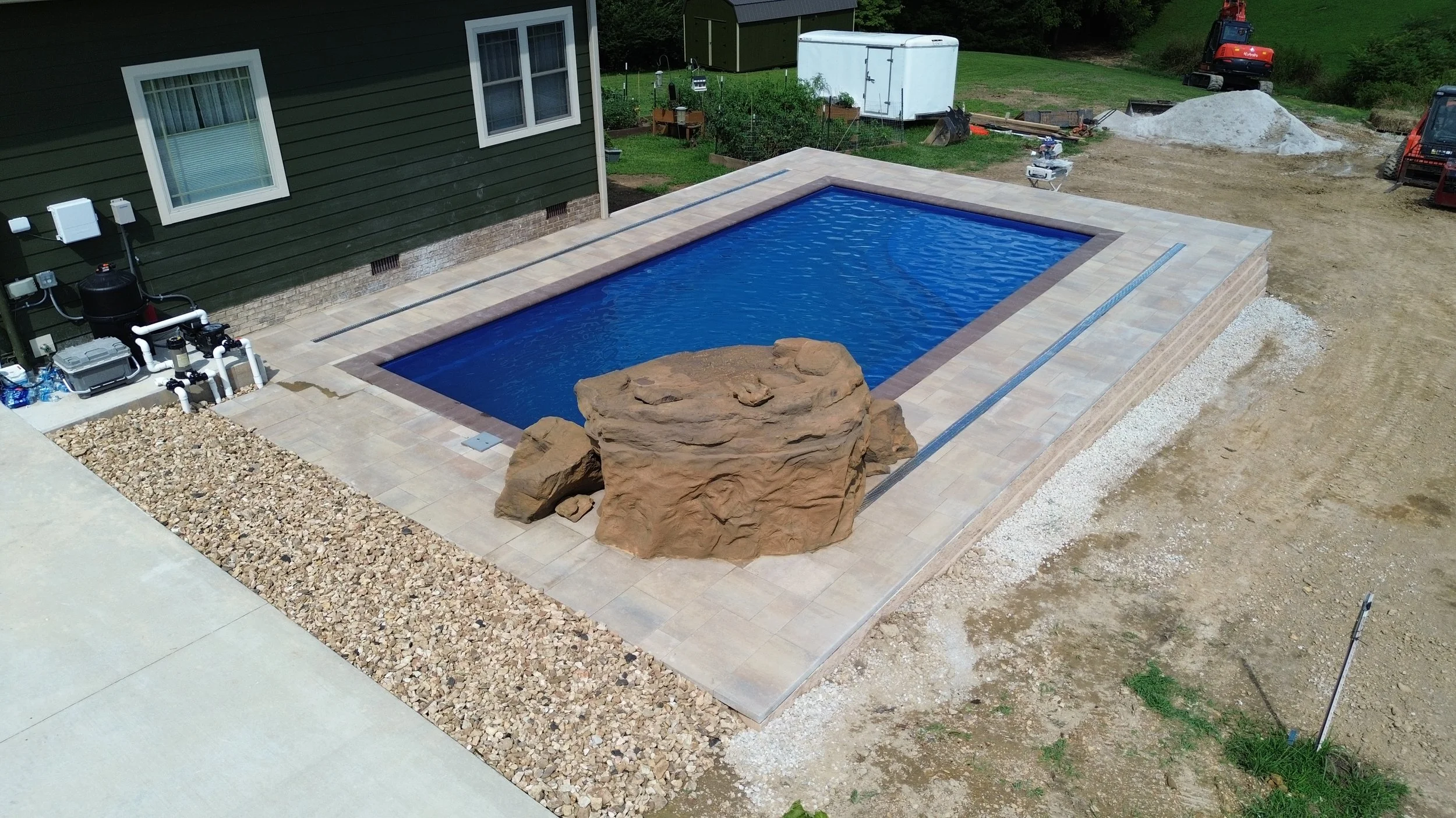 A backyard with a in-ground swimming pool bordered by beige stone tiles. There is a large rock on the edge of the pool, and the area is under construction with exposed dirt and construction equipment in the background.