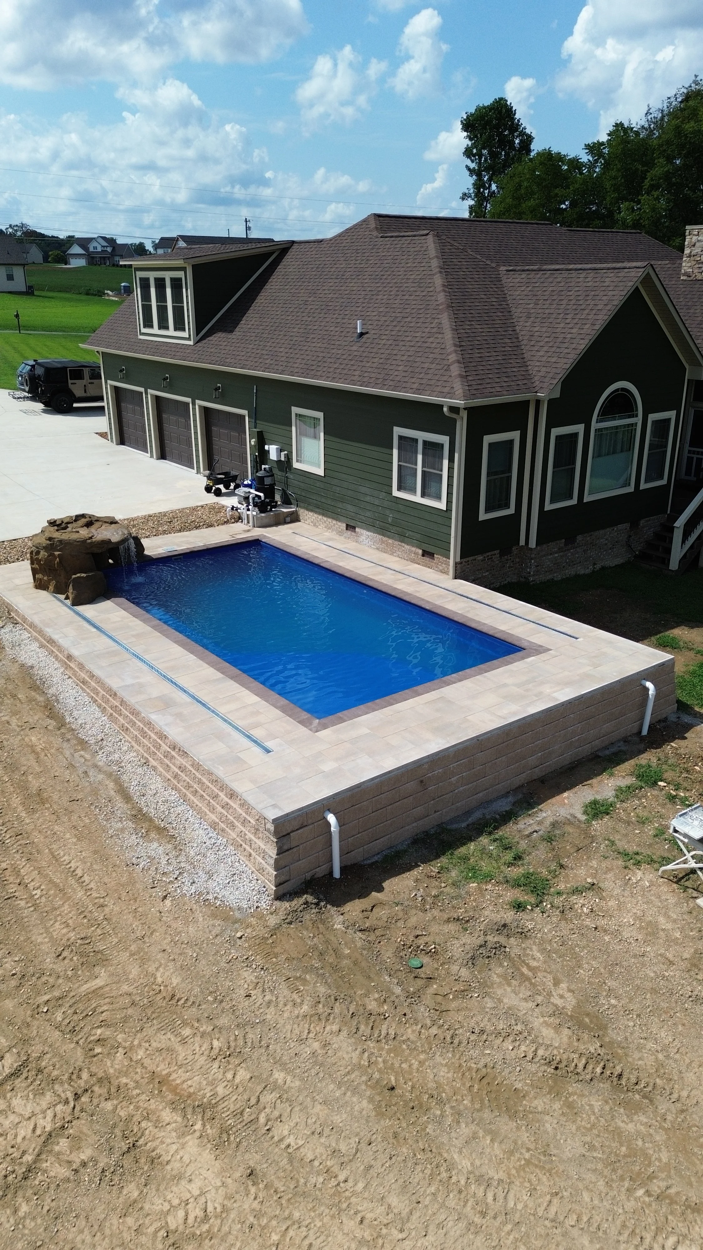 Photo of a backyard with a rectangular in-ground swimming pool with blue water, surrounded by beige paving stones. 