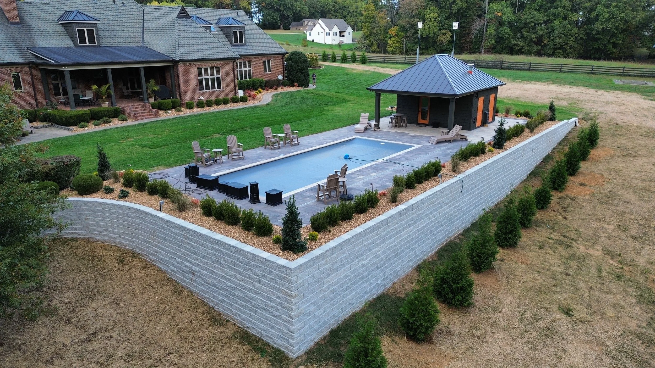 A backyard with a large brick house, a smaller black shed, a swimming pool, outdoor seating, a grassy yard, and a stone retaining wall with landscaping, trees, and neighboring houses in the background.