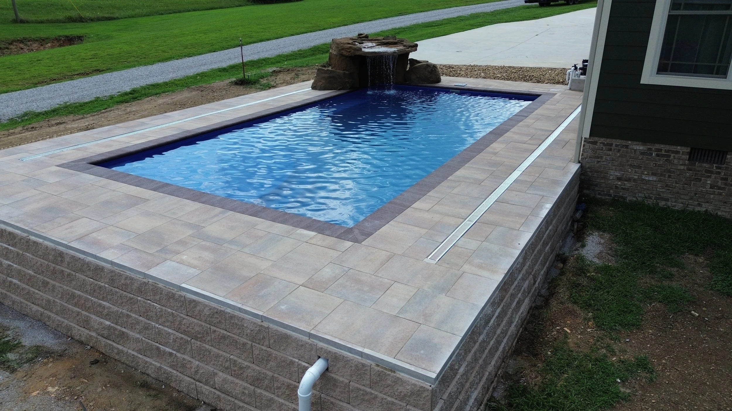 A backyard with a rectangular in-ground swimming pool featuring a rock waterfall and a tiled deck with a drain system, next to a house with siding and a window, beside a grassy lawn and gravel path.