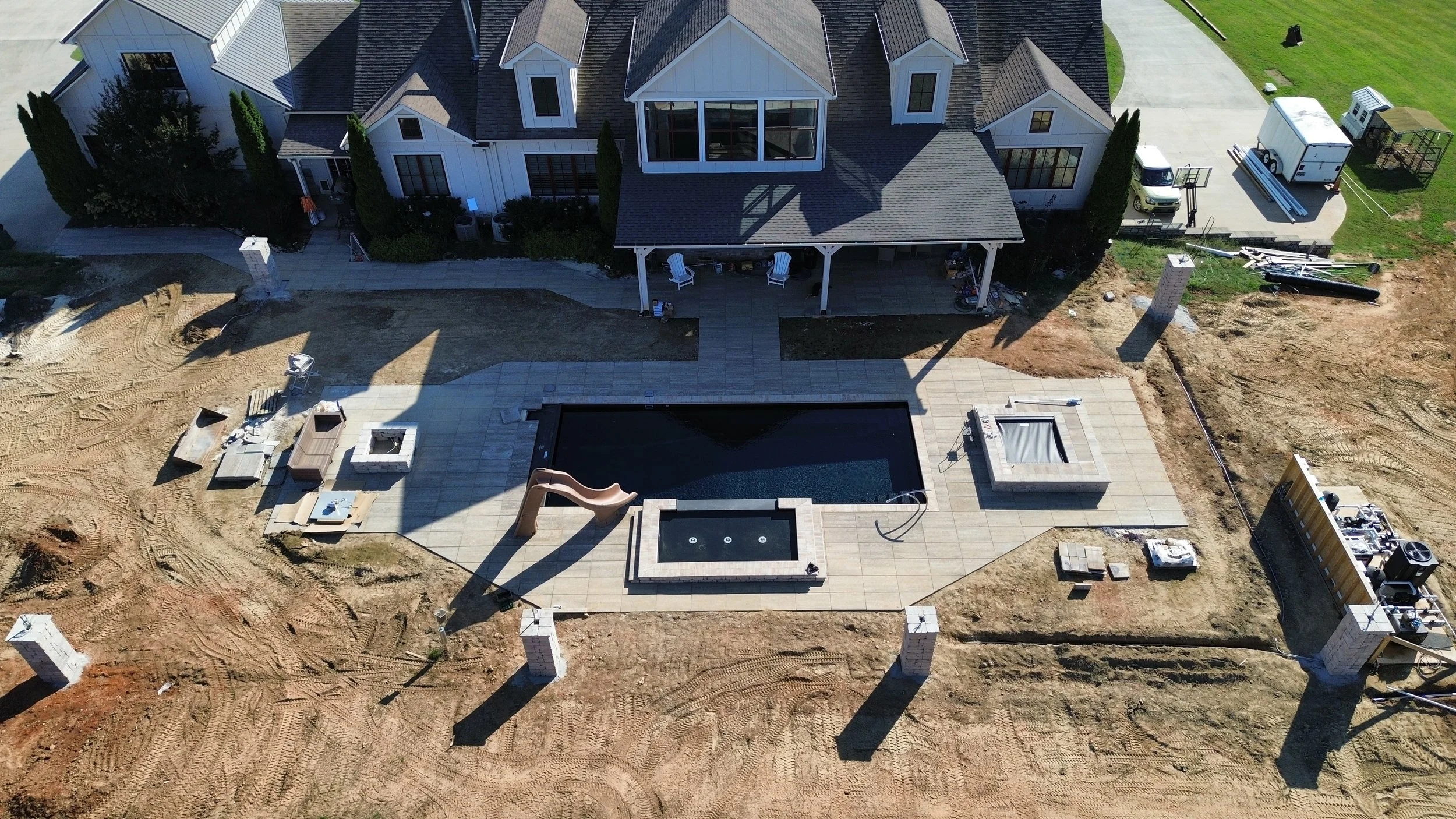 Aerial view of a house under construction with a backyard featuring a pool, hot tub, and playground slide, surrounded by construction materials and equipment.