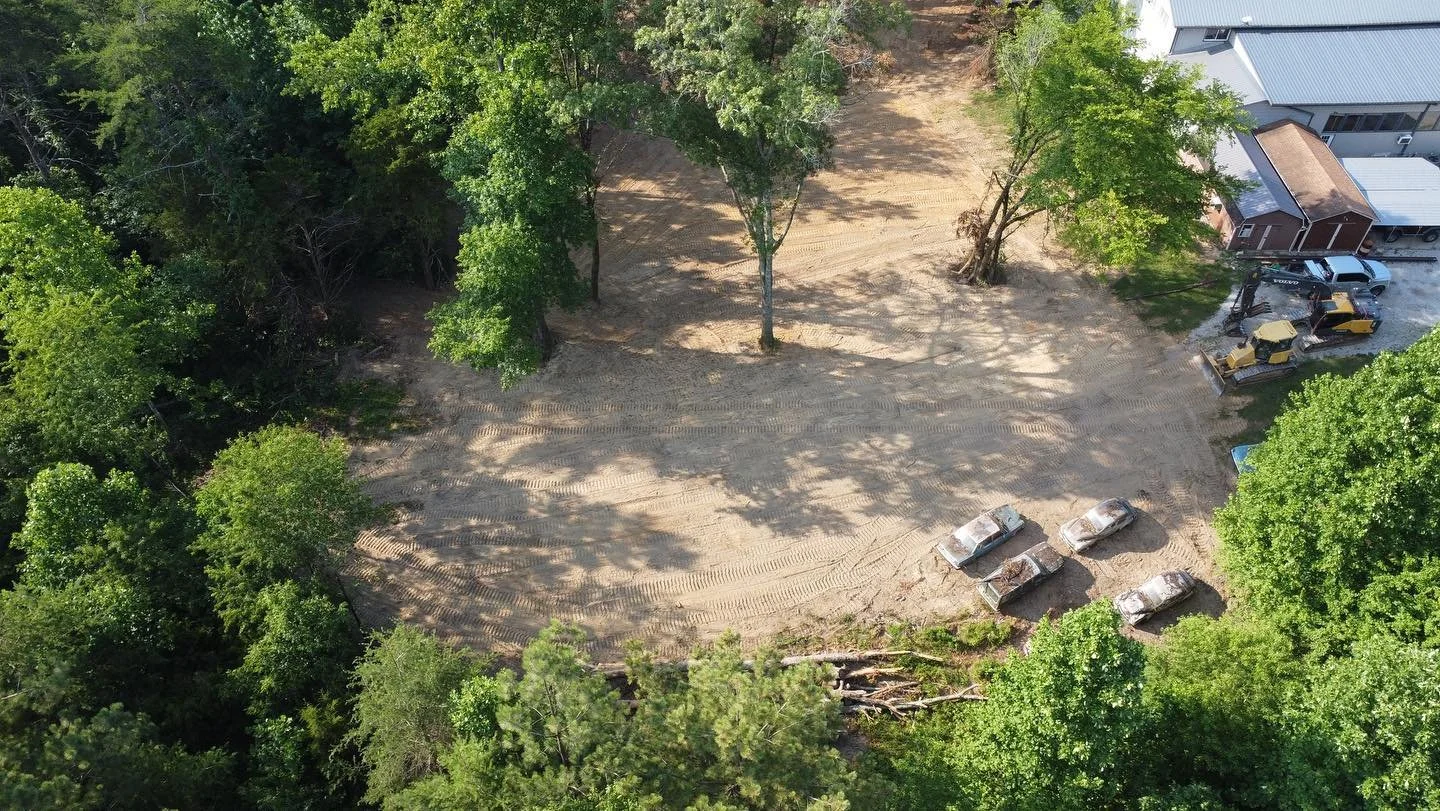 An aerial view of a cleared lot with five burned cars parked in a row, surrounded by green trees. There is construction equipment and a house on the right side.
