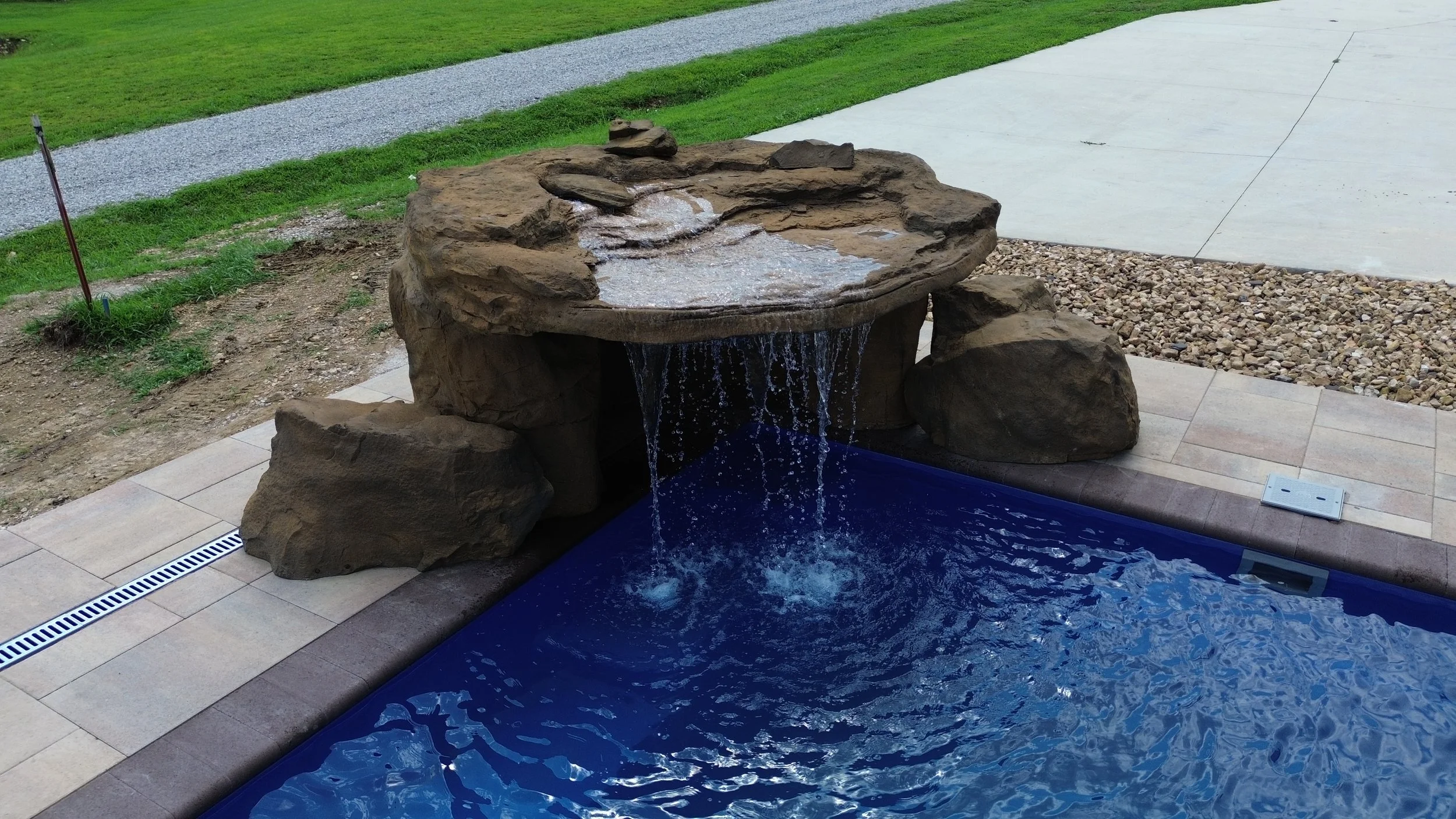 Artificial rock fountain with water flowing from top into a small pool, set on a backyard patio with grass, gravel, and concrete area in the background.