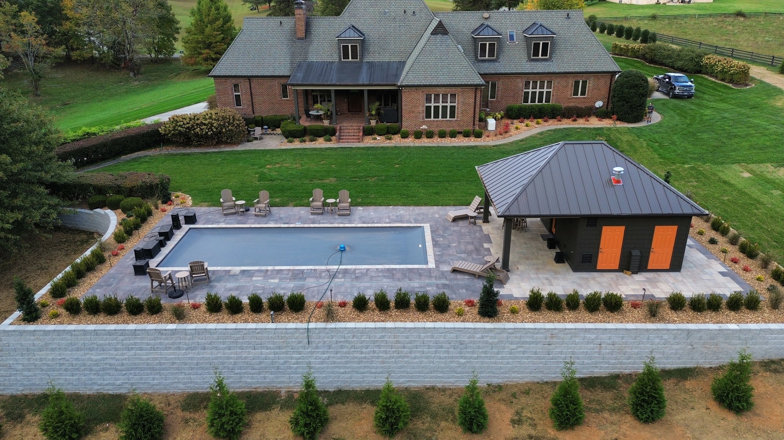 Aerial view of a backyard with a swimming pool, patio furniture, a small black pool house with orange doors, a well-manicured lawn, and a large brick house with multiple windows and a gray roof surrounded by trees and landscaping.
