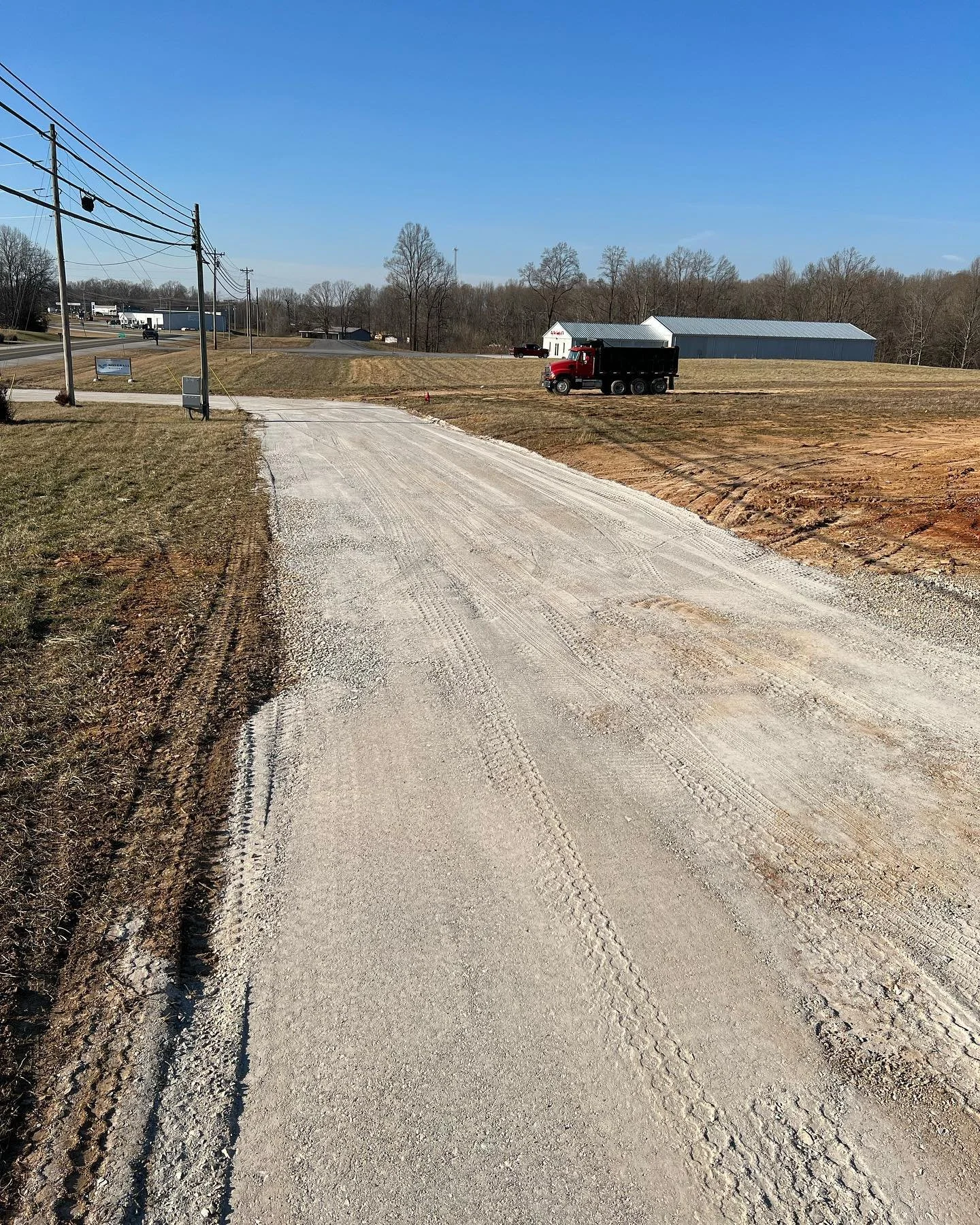 A gravel driveway leading to a grassy area with a red truck parked near a large gray building with a metal roof, trees in the background, power lines running along the left side, and a blue sky overhead.