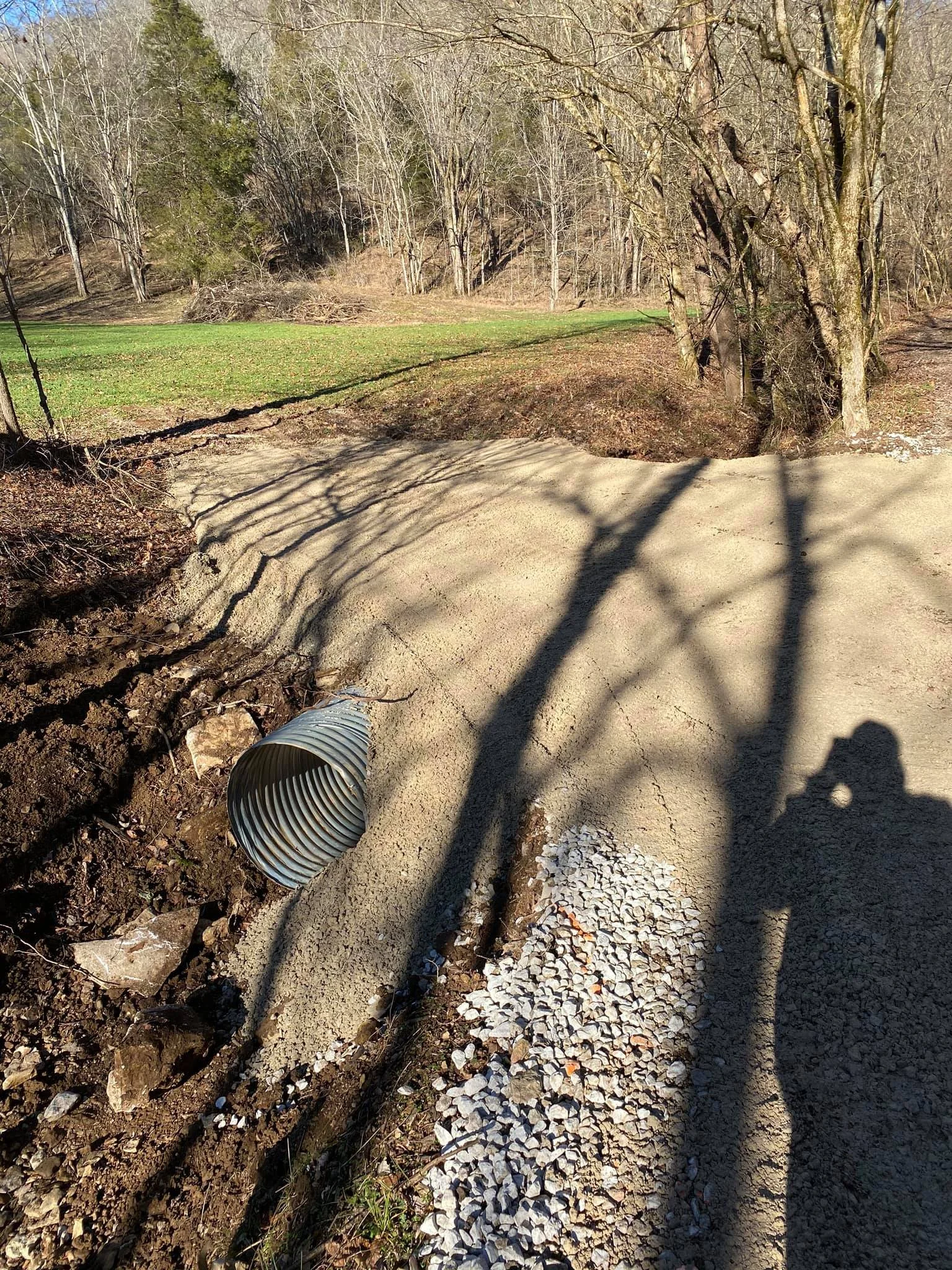 A metal drainage pipe alongside a gravel pathway in a wooded area with leafless trees and grassy field.