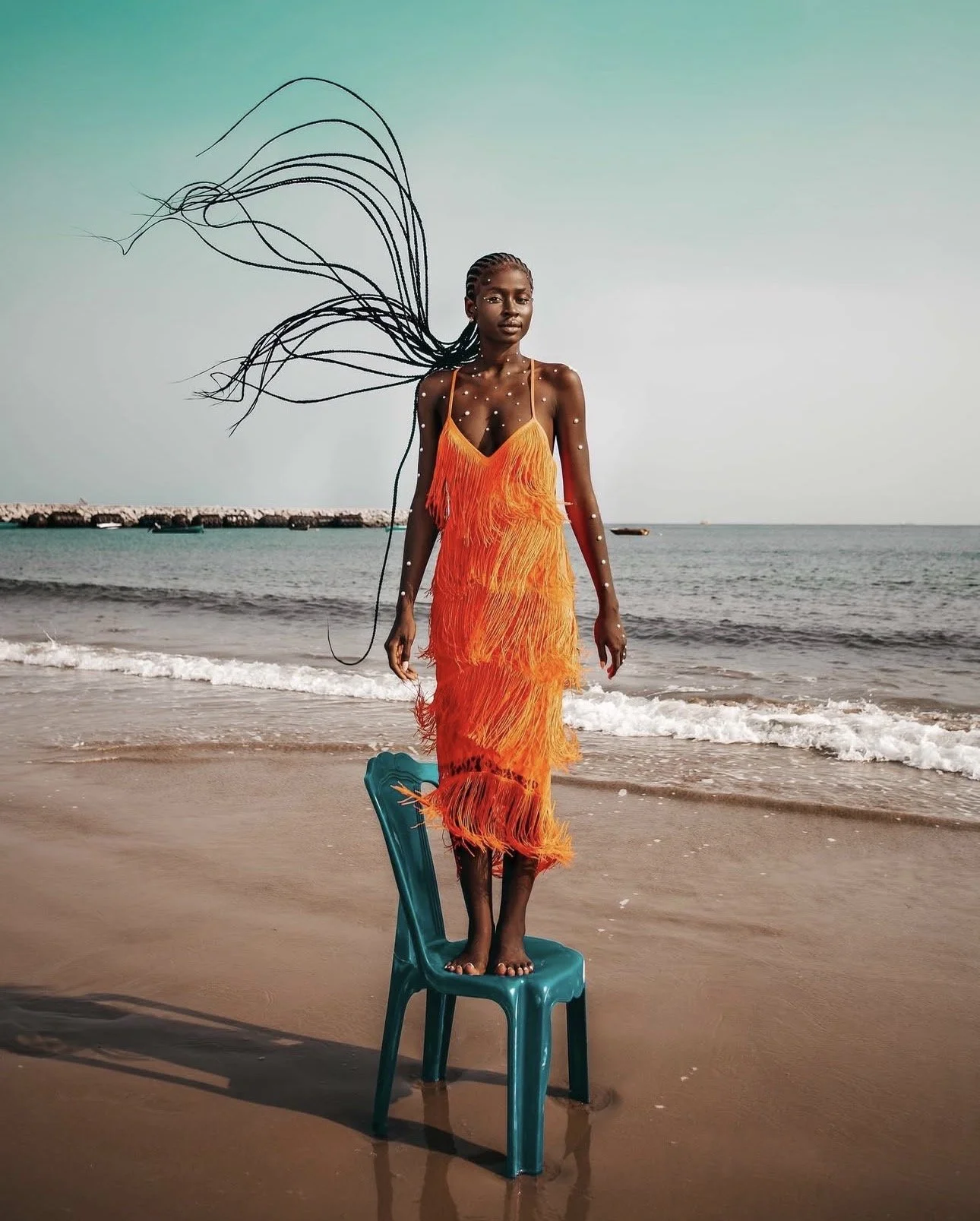 A woman standing on a plastic chair on a sandy beach, wearing a bright orange dress with fringe, with her hair flowing in the wind and ocean waves in the background.