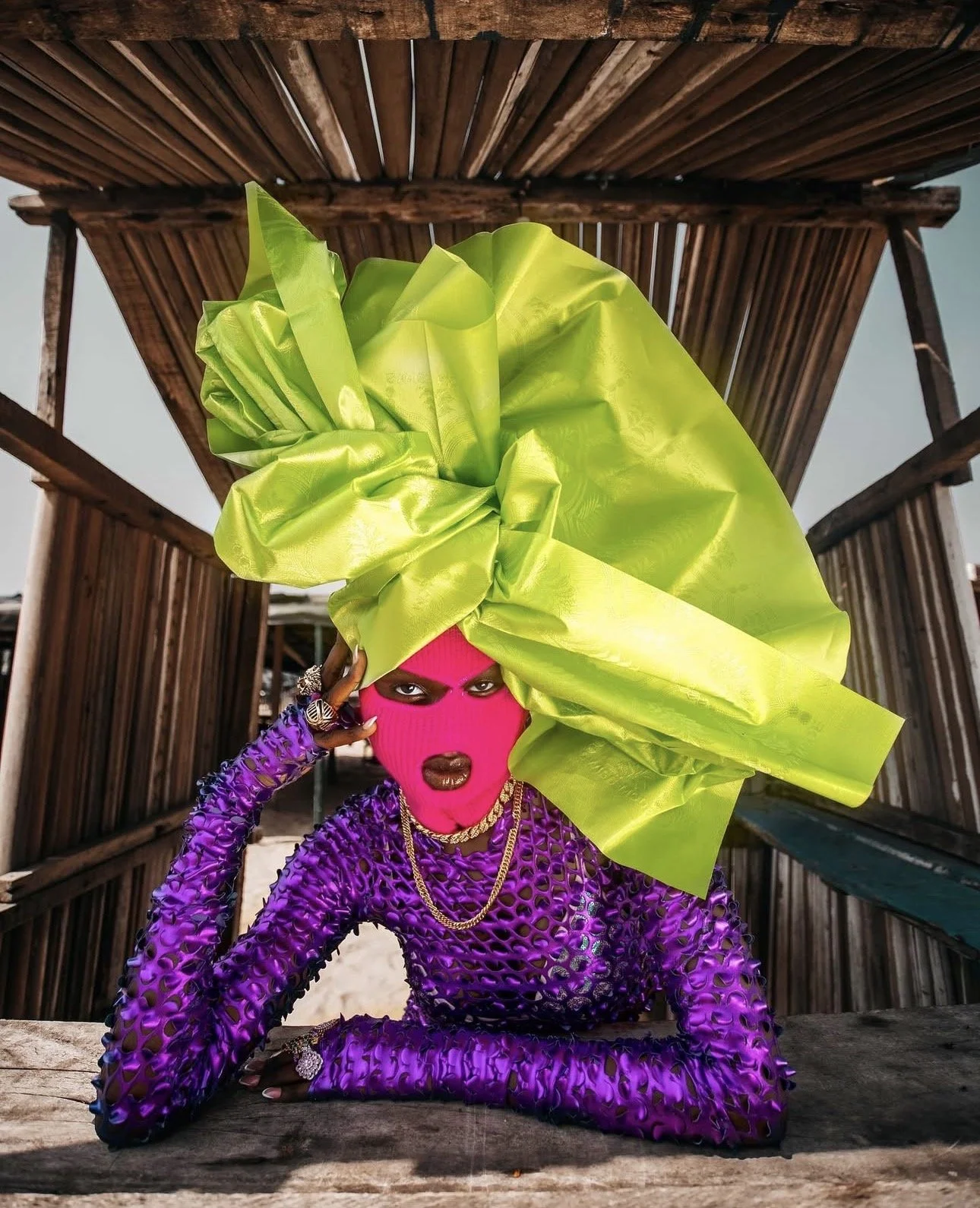 A person wearing a pink ski mask, dark lipstick, and purple fishnet outfit with jewelry, under a wooden structure, with a large, bright green bow on their head.