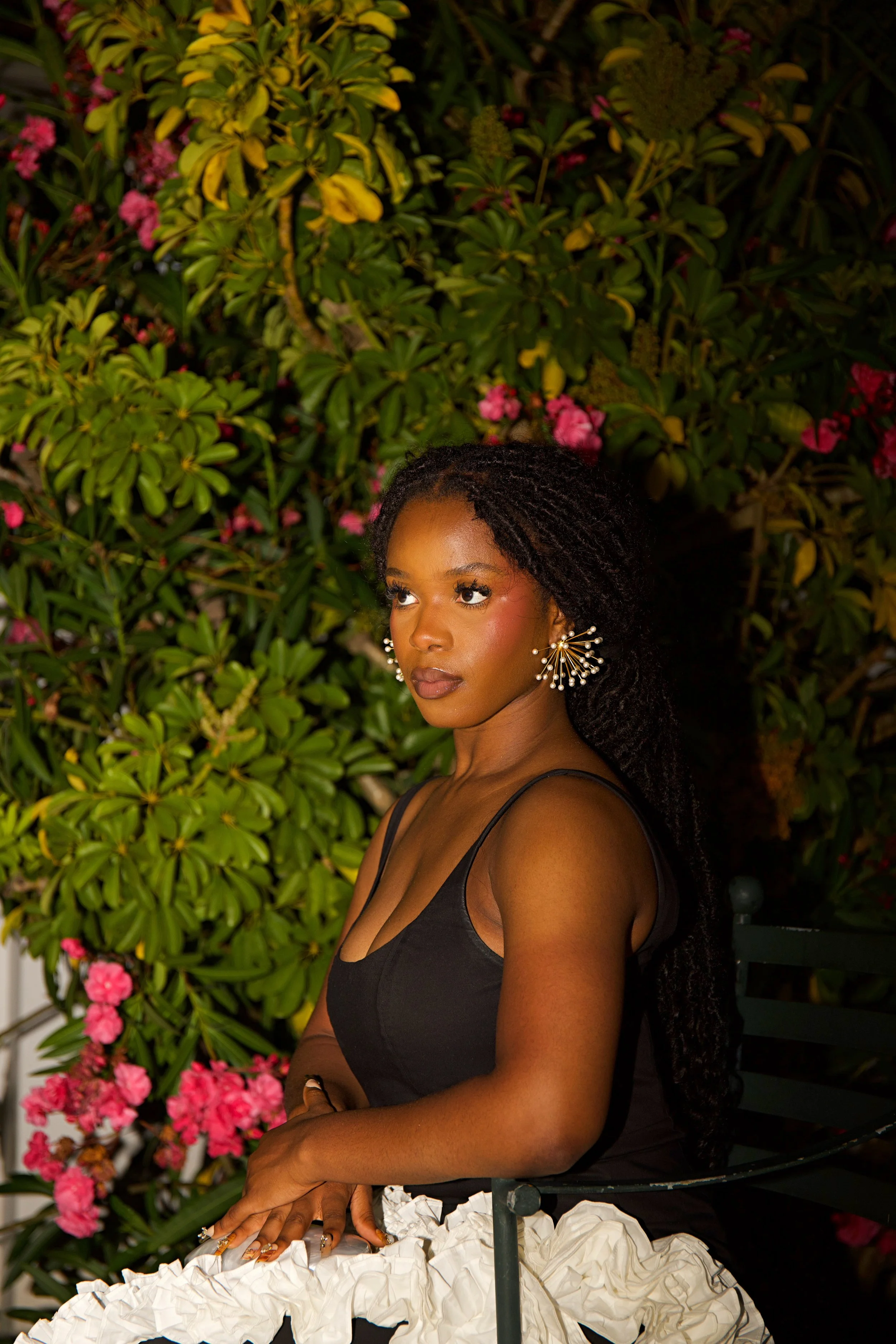 A woman with dark skin and long braided hair, wearing a black top and white ruffled skirt, sitting outdoors against a background of large green bushy plants with pink flowers.