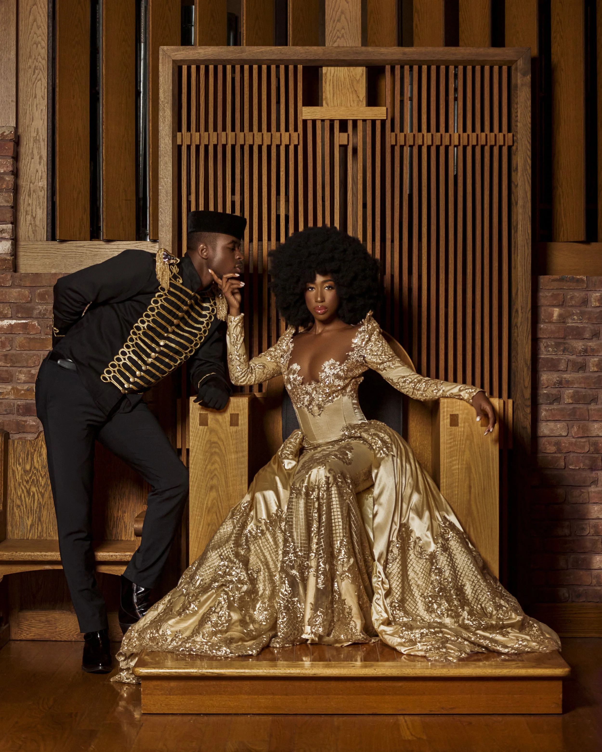 A woman dressed in an elaborate gold gown sitting on a wooden throne, with a man dressed in a black military-style uniform with gold accents standing next to her, in a room with brick and wood walls.