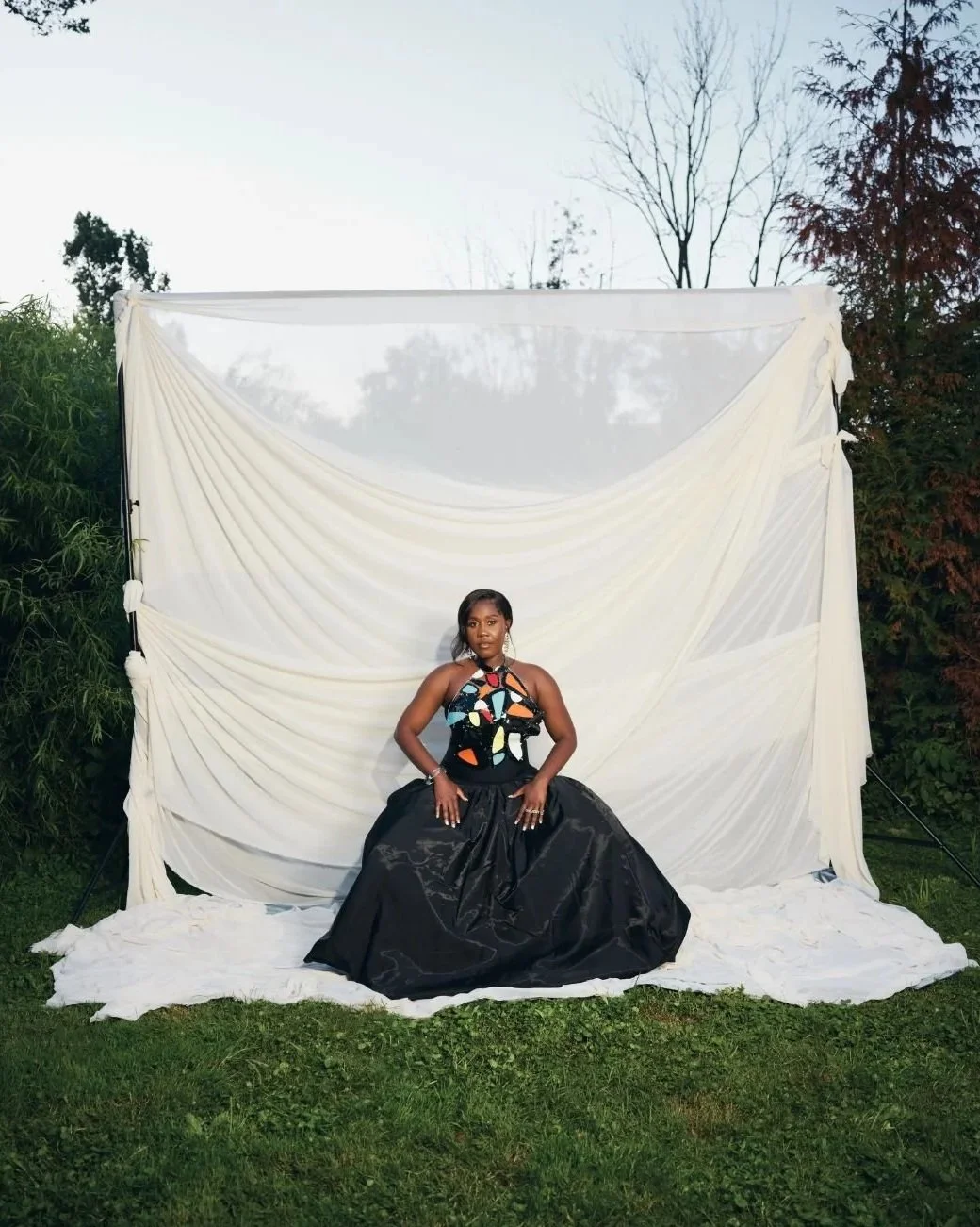 A woman in a colorful, patterned top and a long black satin skirt seated on a white fabric-covered surface outdoors in front of a white draped backdrop, with trees and sky in the background.
