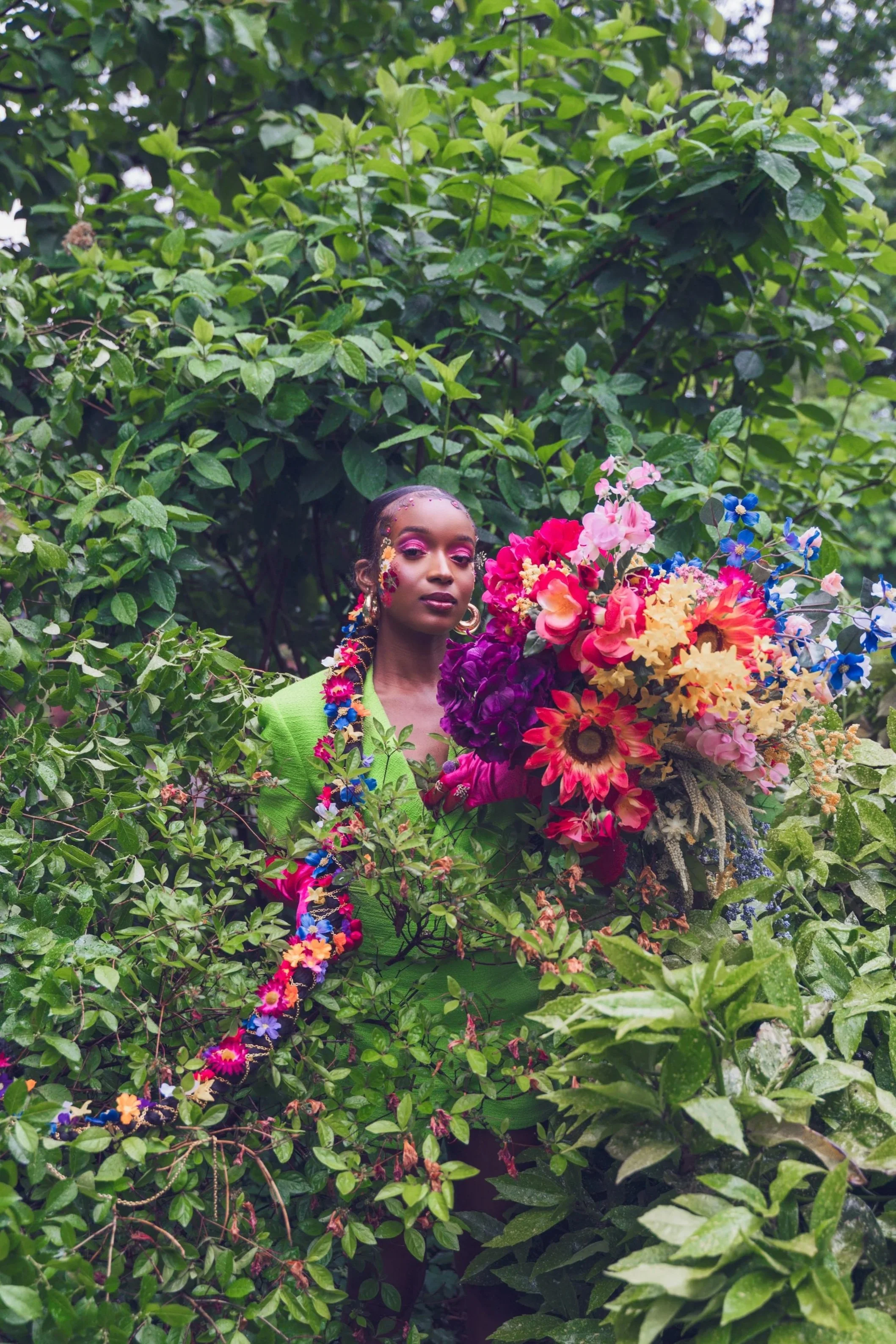 A woman with decorated face and braided hair, wearing a bright green jacket, standing amidst lush green foliage, holding a large colorful bouquet of flowers.
