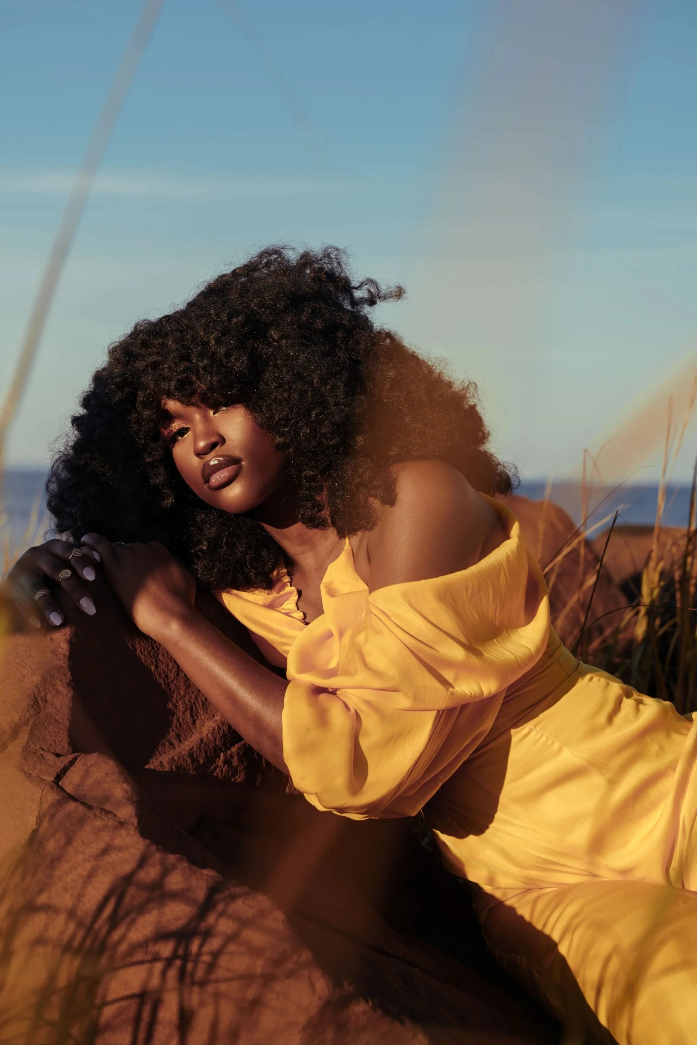 A woman with curly black hair posing outdoors in a yellow dress during sunset.