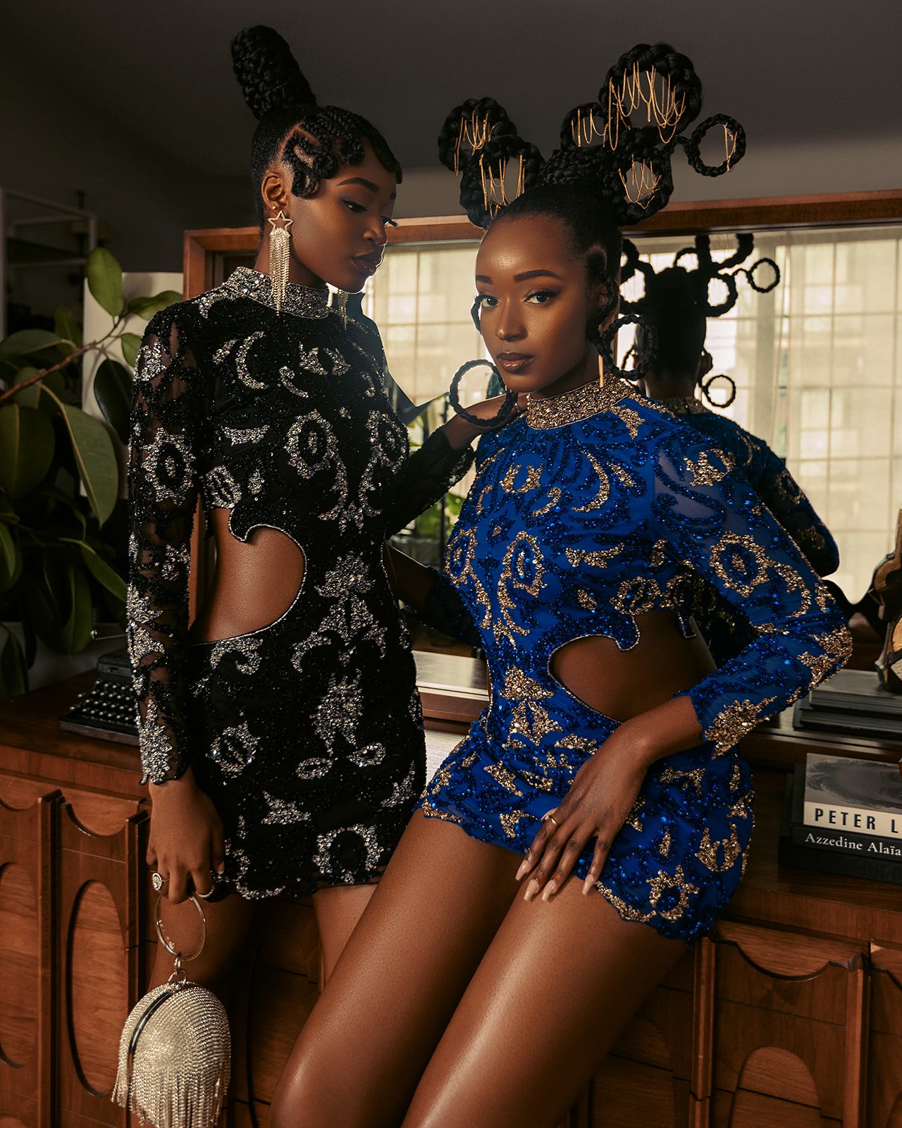 Two women with elaborate braided hairstyles wearing embellished cutout dresses in black and blue, posing indoors near a wooden cabinet and window.