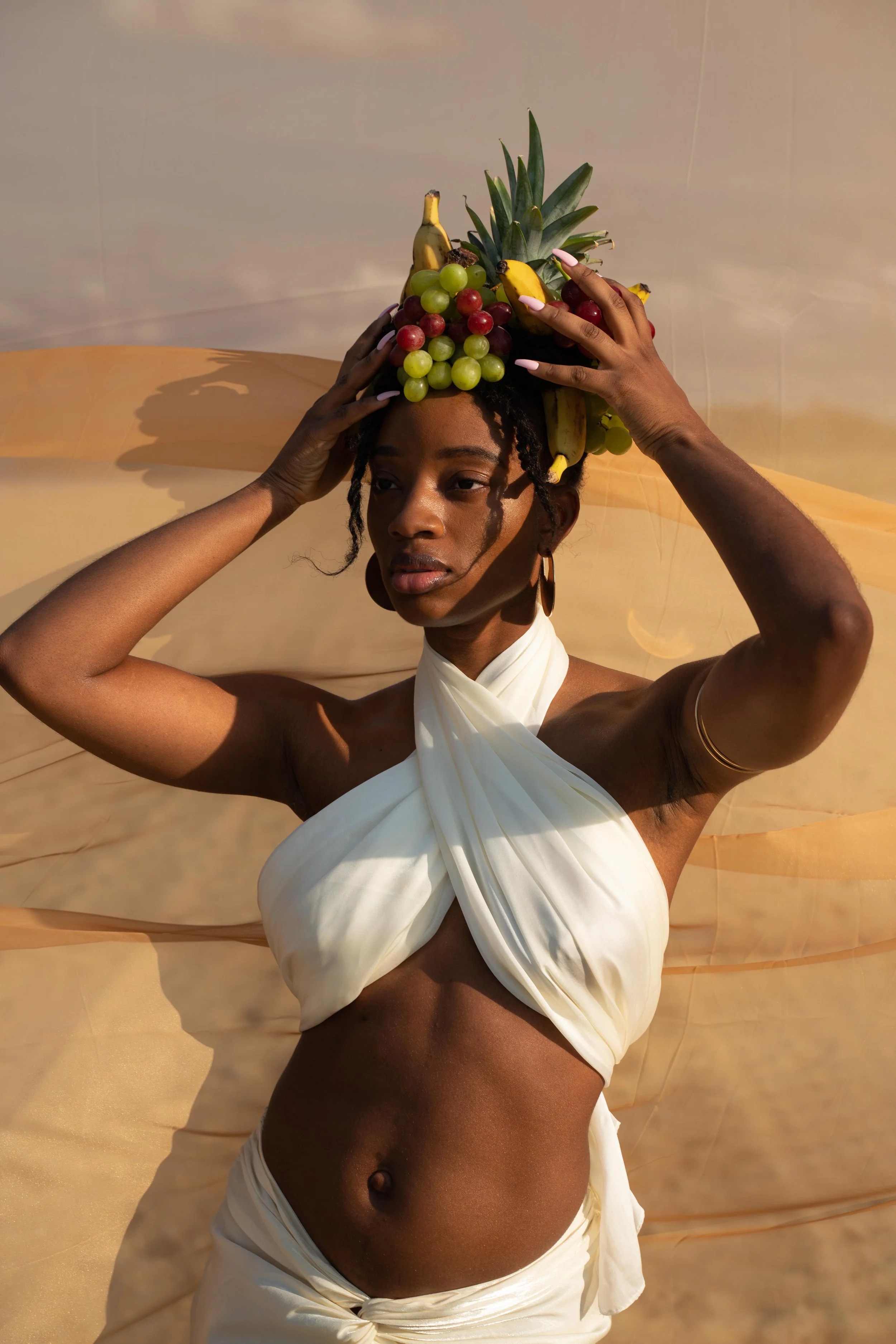 A woman with dark skin and curly hair styled in twists, wearing a white, draped top and a matching skirt, balancing a throne-like arrangement of various fruits, including a pineapple, bananas, grapes, and other tropical fruits, on her head, with a ne