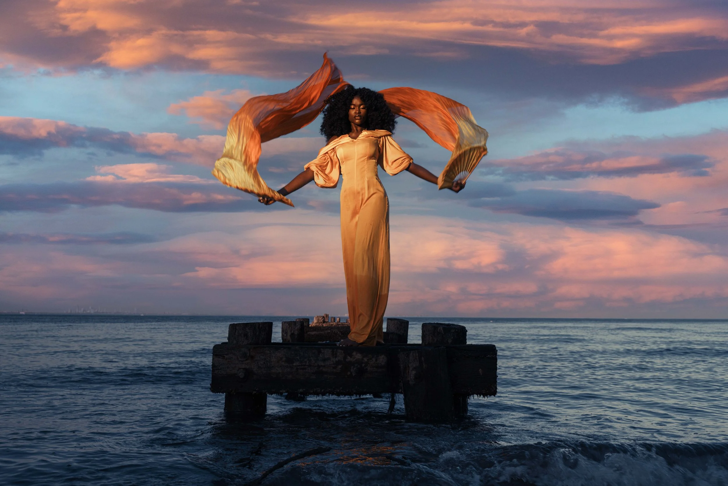 A woman in a golden dress standing on a wooden platform in the ocean during sunset, holding a flowing fabric above her head.