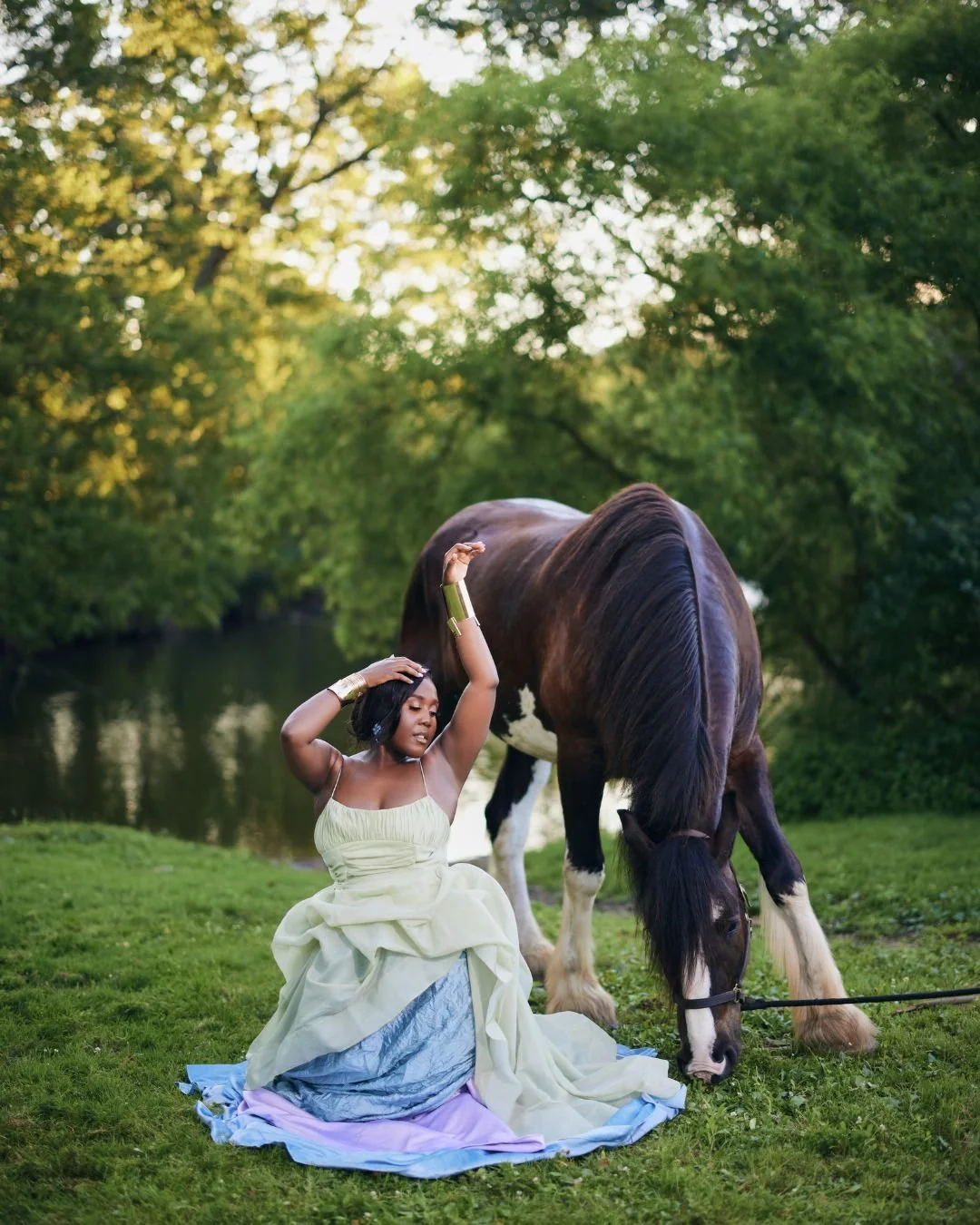 A woman in a flowing yellow dress with a lilac and blue skirt sitting on the grass near a pond, touching her hair, with a large brown and white horse grazing nearby, surrounded by lush green trees.