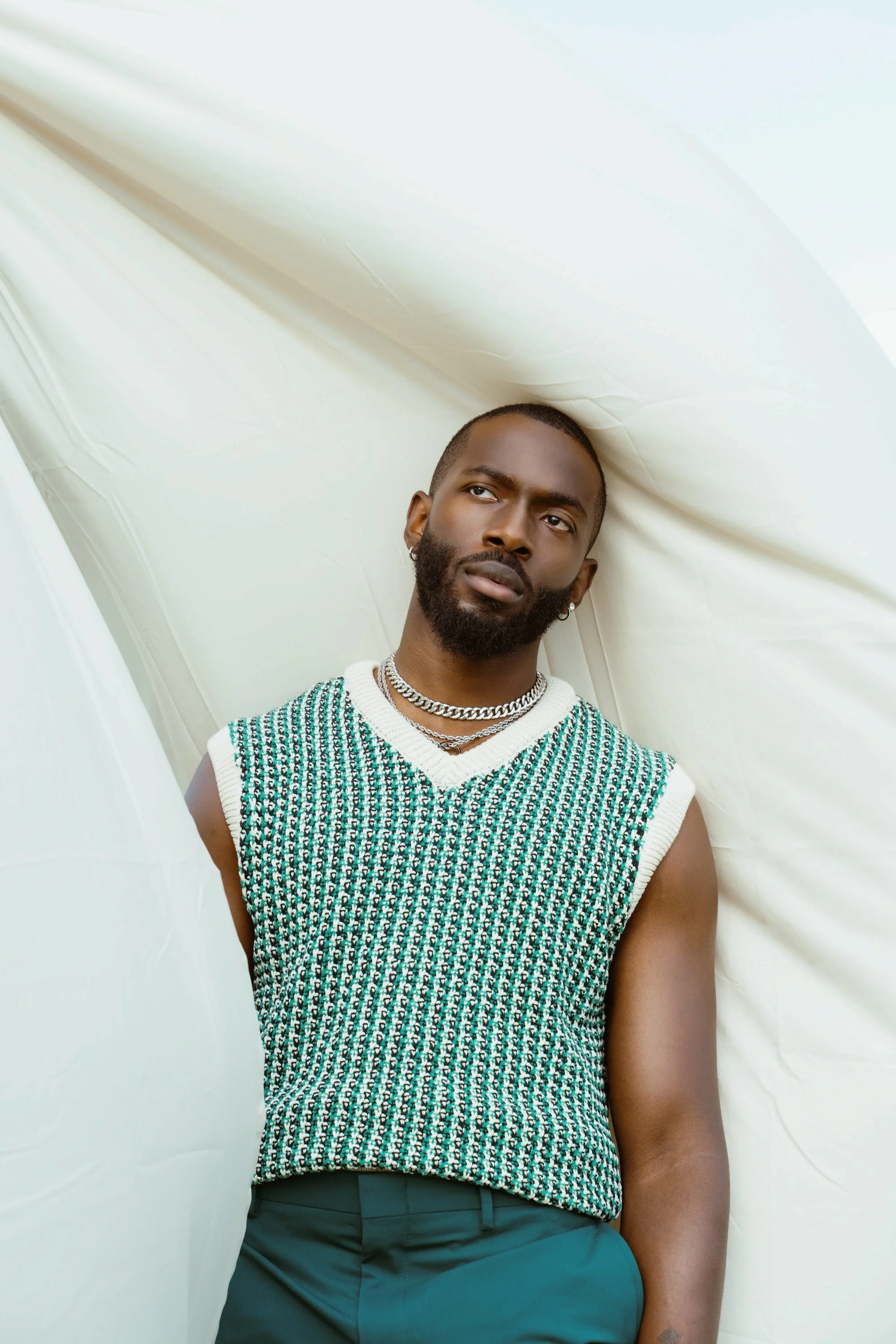 A man with a beard and short hair wearing a patterned sleeveless sweater, layered necklaces, and teal pants, standing against white fabric background.