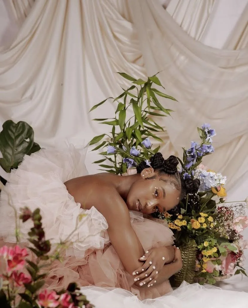 A woman lying down amidst a lush arrangement of colorful flowers and green plants, with a backdrop of cream-colored draped fabric.