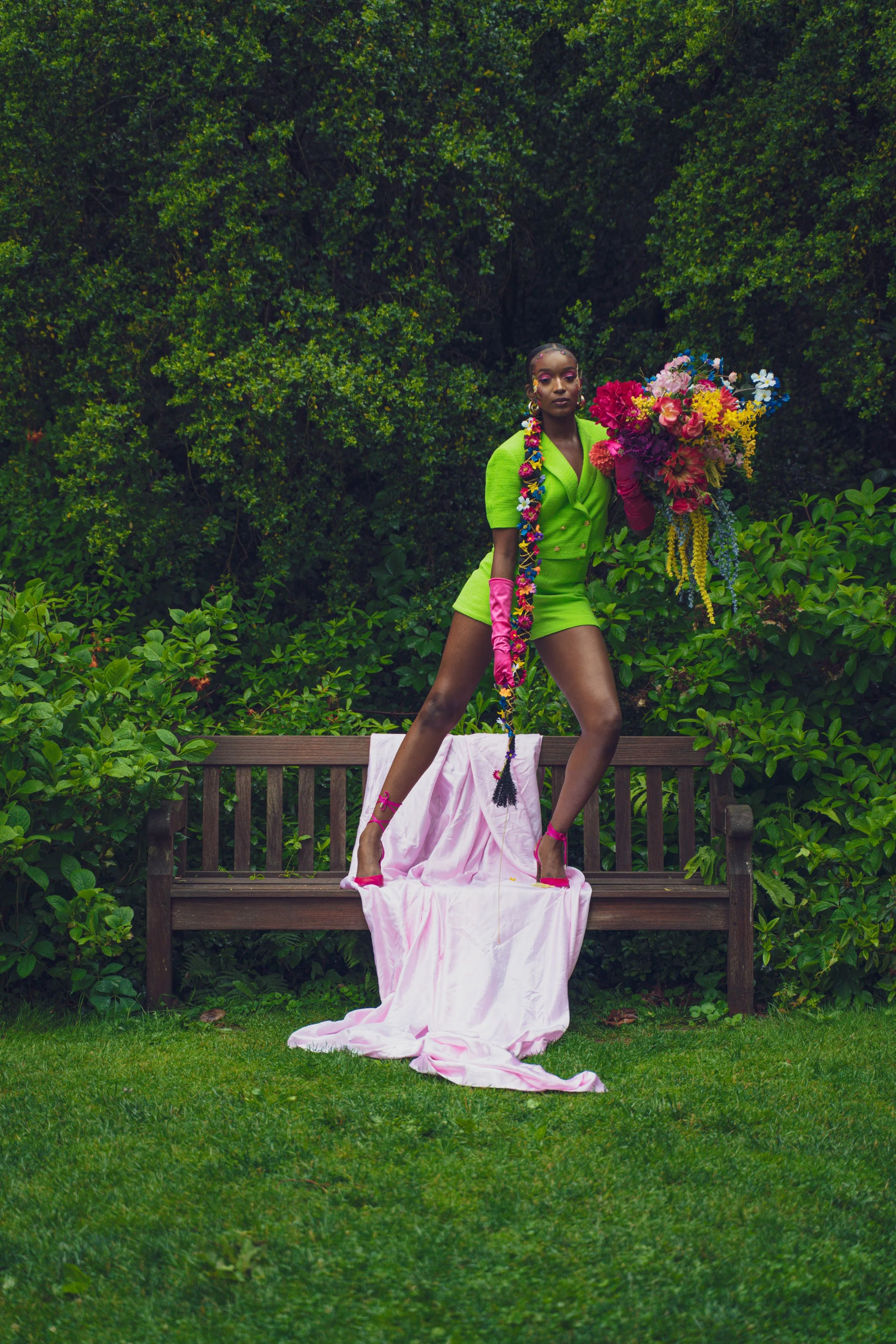 A person standing on a garden bench holding a colorful bouquet of flowers, wearing a bright green outfit, pink gloves and shoes, with a long braided hairstyle decorated with flowers, surrounded by lush green foliage.