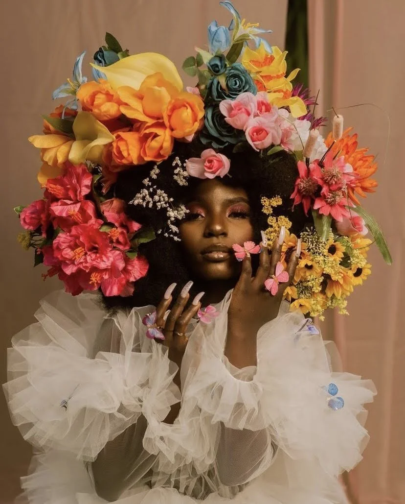 A woman with dark skin wearing a white, ruffled, sheer dress, surrounded by an elaborate headdress made of colorful flowers including roses, daisies, and other blossoms.