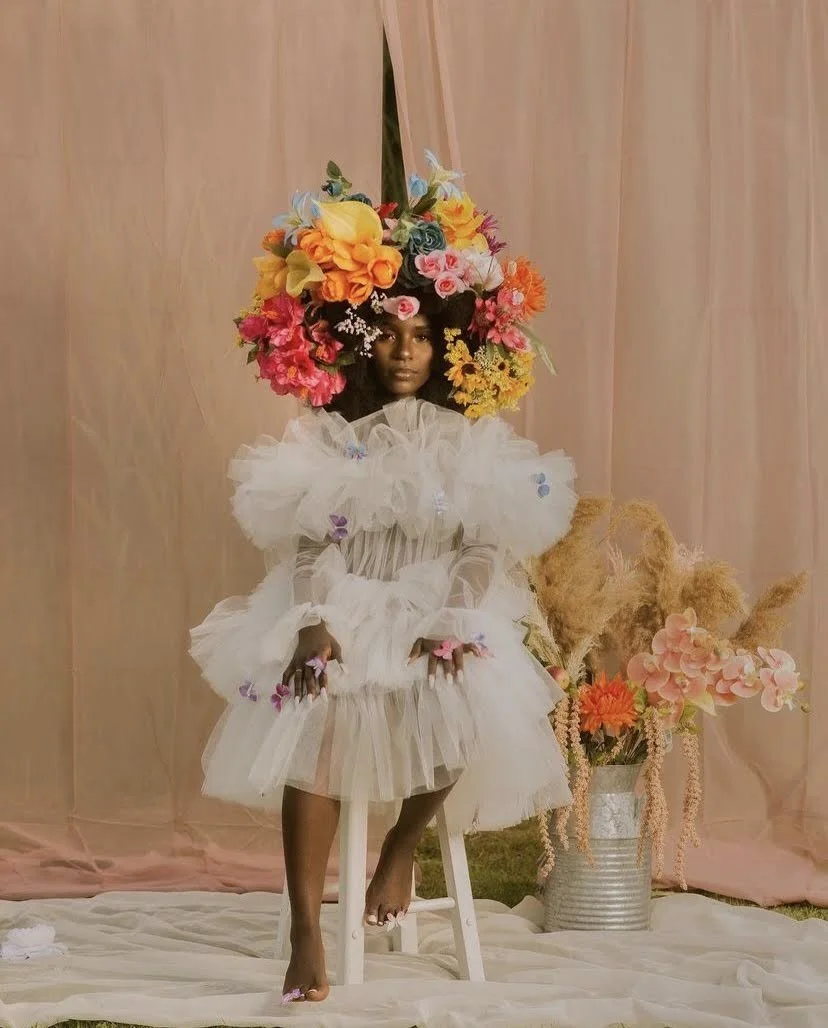 A woman sitting on a stool with a large floral headdress and a fluffy white dress in front of a pink curtain with a vase of flowers beside her.