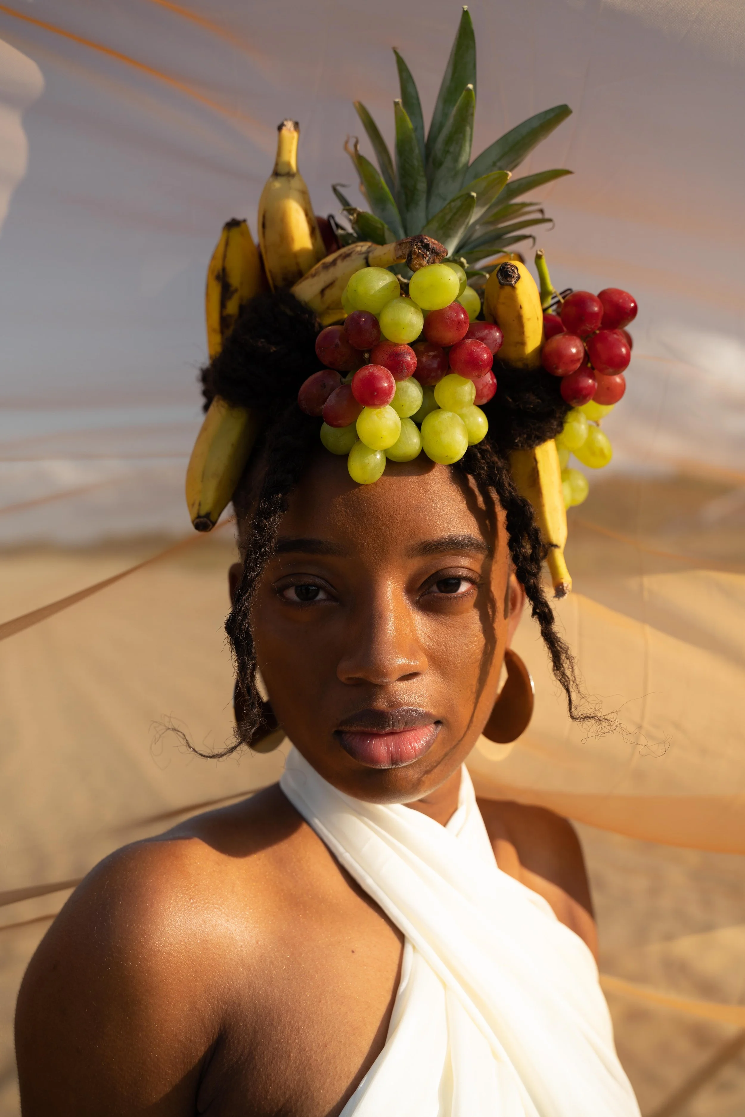 Portrait of a woman with fruit accessories in her hair, featuring bananas, grapes, and a pineapple, wearing a white halter top, against a warm, blurred background.