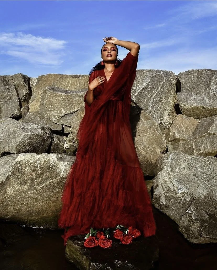 A woman in a flowing red gown stands against a rocky shoreline with blue sky overhead, holding one hand to her forehead and the other near her chest, with red roses at her feet.