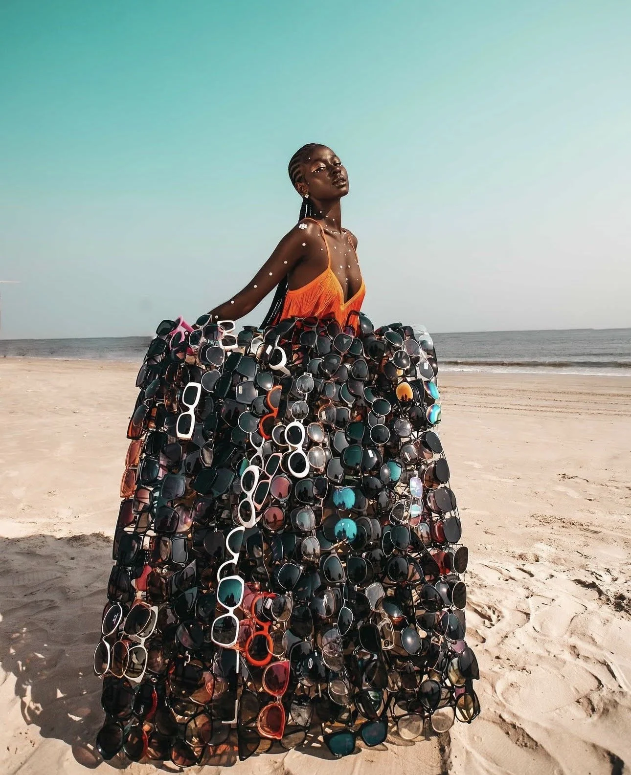 A woman standing on a sandy beach wearing a fringe dress standing behind a board of numerous sunglasses with a ocean in the background.