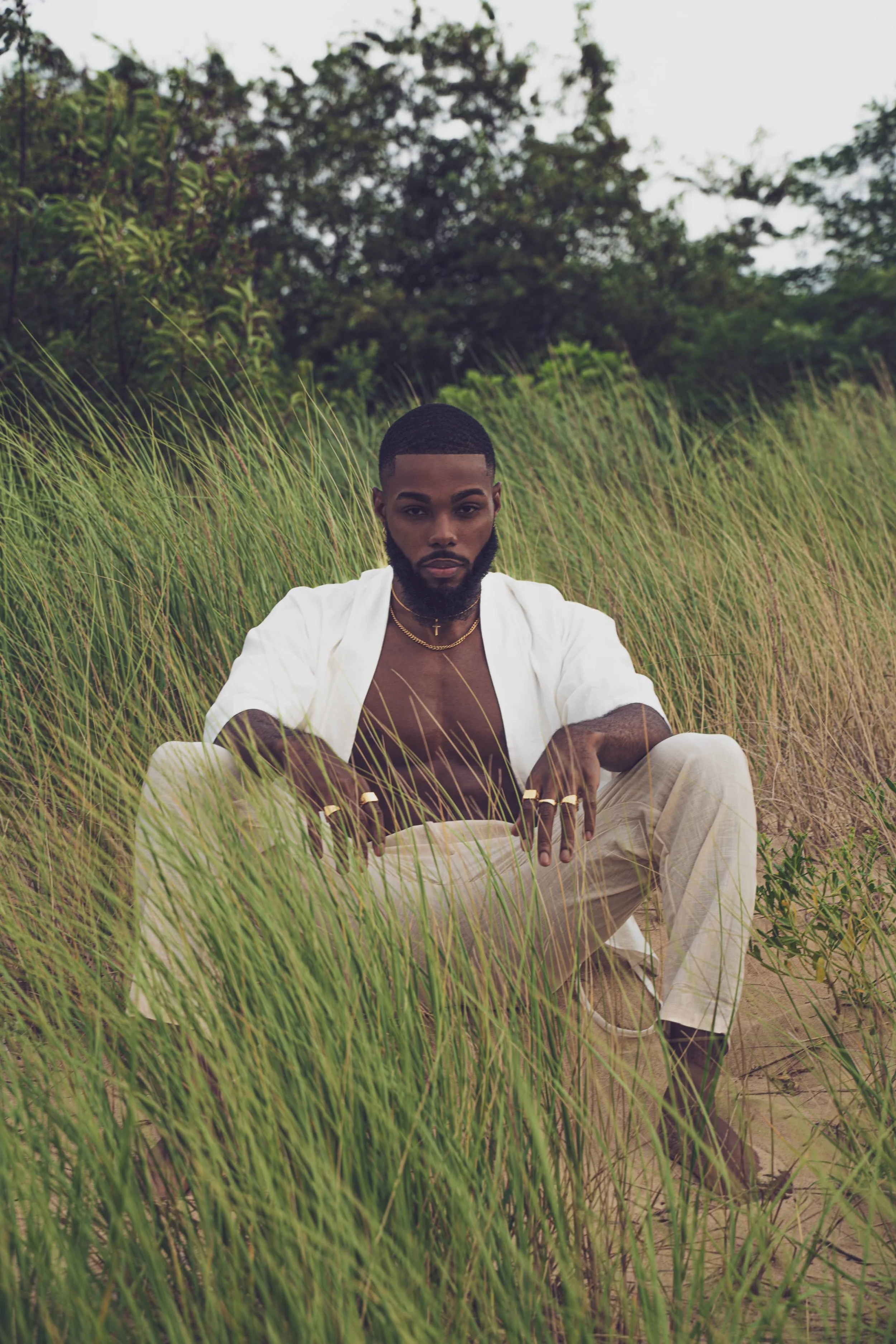 A man crouching in tall grass outdoors, wearing beige pants, a partially open white shirt, a gold necklace, and rings, with trees in the background.