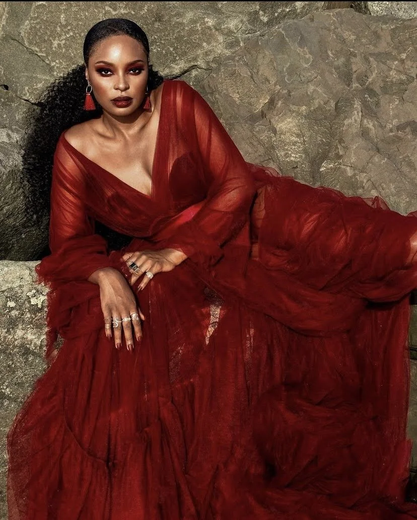 A woman with dark curly hair and dark lipstick, wearing a sheer red dress with a deep neckline, layered with a matching red tulle skirt, seated against a rocky background, accessorized with earrings and rings.